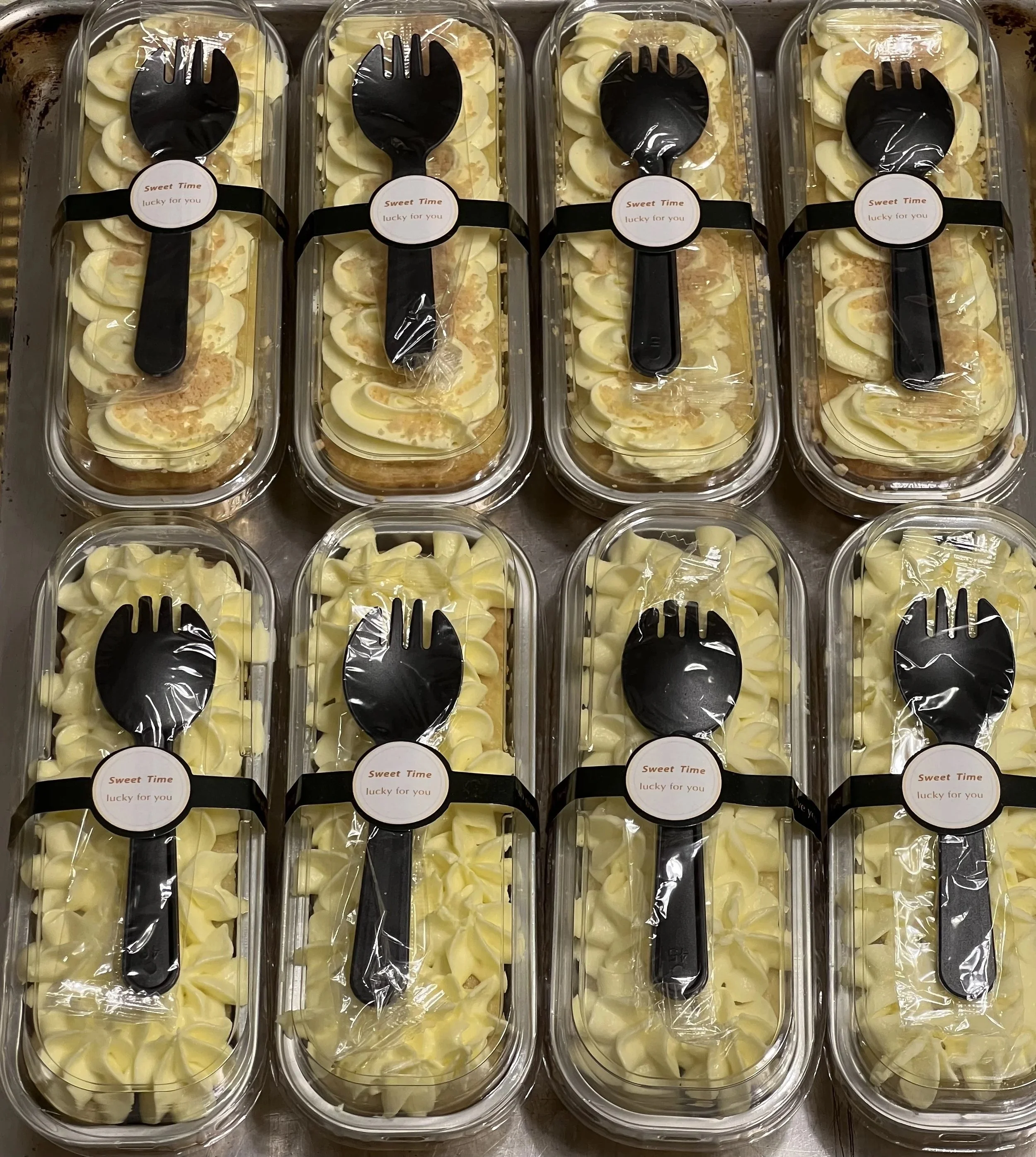 Eight clear plastic containers filled with sliced bananas topped with whipped cream, each sealed with a plastic wrap and a black spoon attached with a sticker labeled 'Sweet Time lucky for you'.