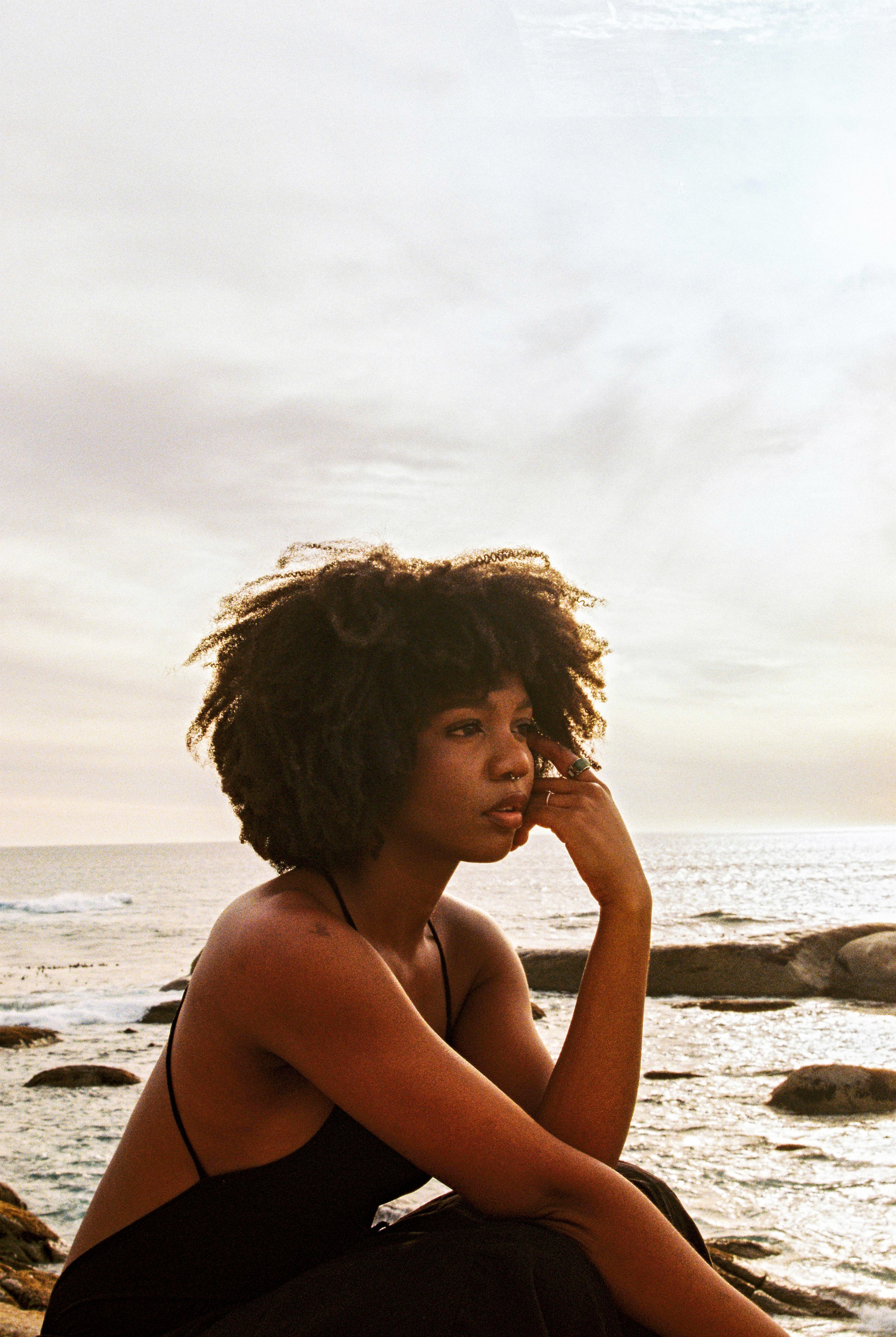 A young woman with curly hair sitting on rocks by the ocean at sunset, gazing into the distance with her hand resting on her face.