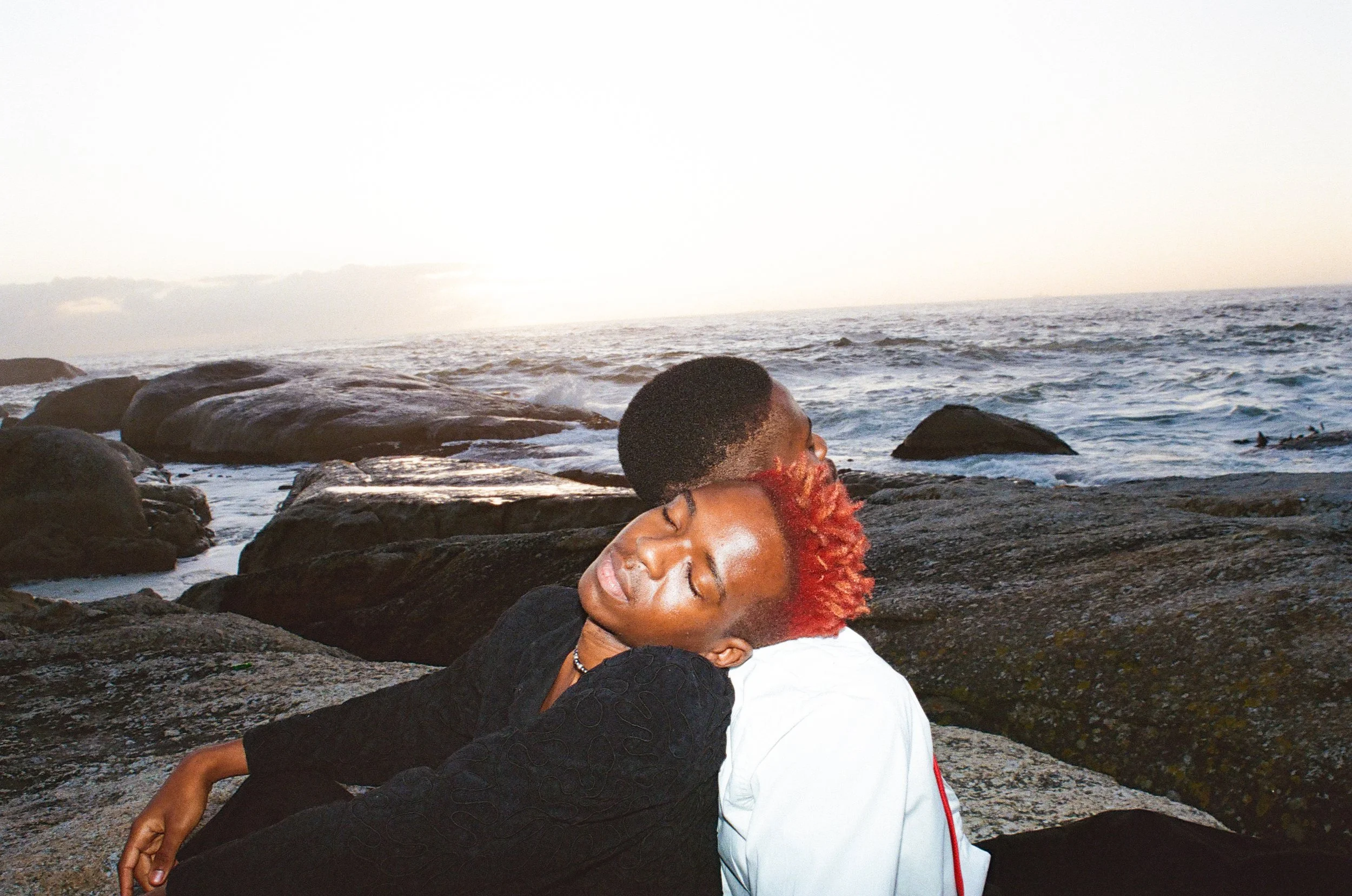 Dnemas and a friend with closed eyes resting against each other on rocks by the ocean at sunset, with waves and a cloudy sky in the background.