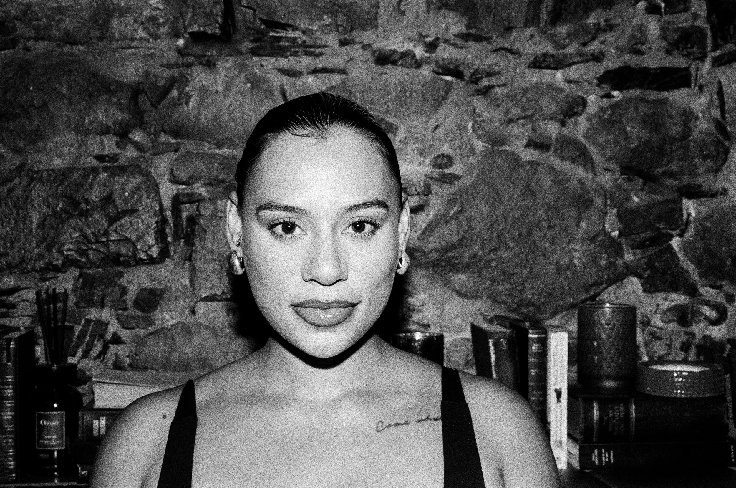 Kimberleigh Venty with dark hair in an updo, wearing earrings and a sleeveless top, standing in front of a stone wall with a bookshelf containing books and decorative items.