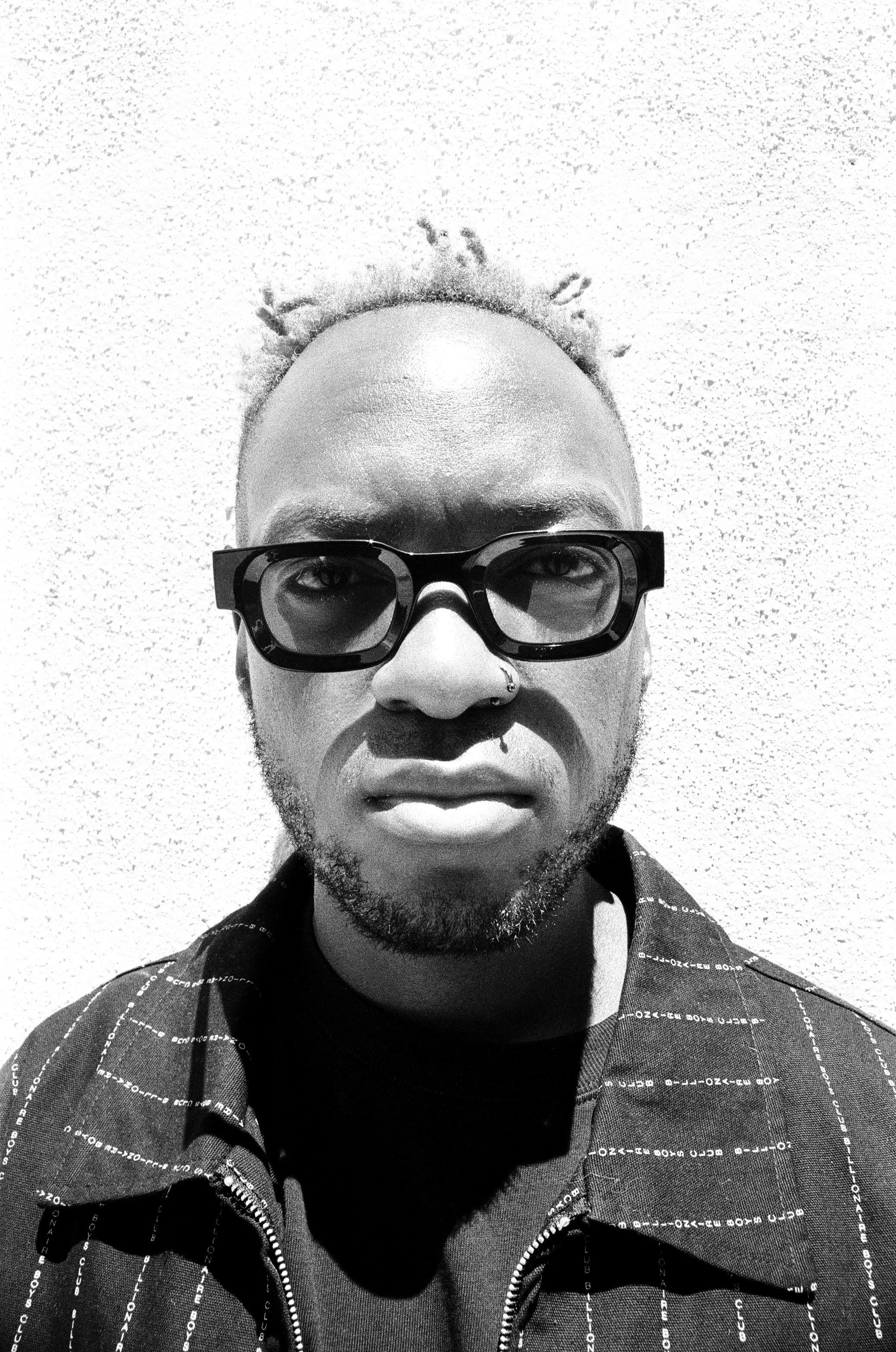 Black and white close-up portrait of Themba Hadebe, short dreadlocks, wearing thick black sunglasses, a nose ring, and a zip-up jacket with printed text.