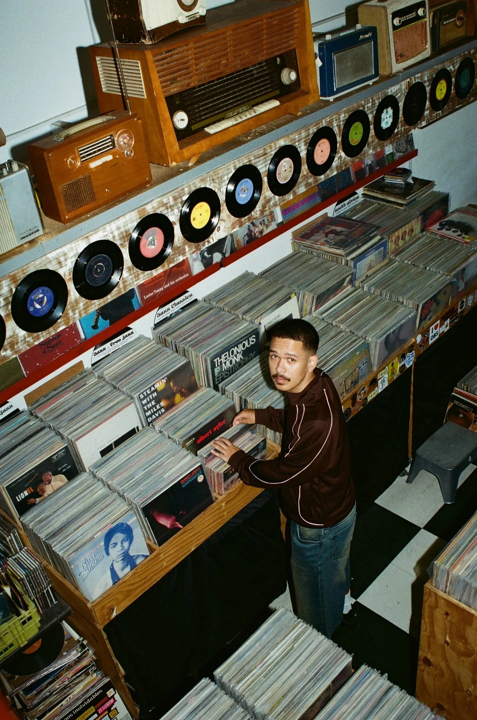 Jarry Pitboi browsing vinyl records in a record store with shelves of records and vintage radios, black and white checkered floor.