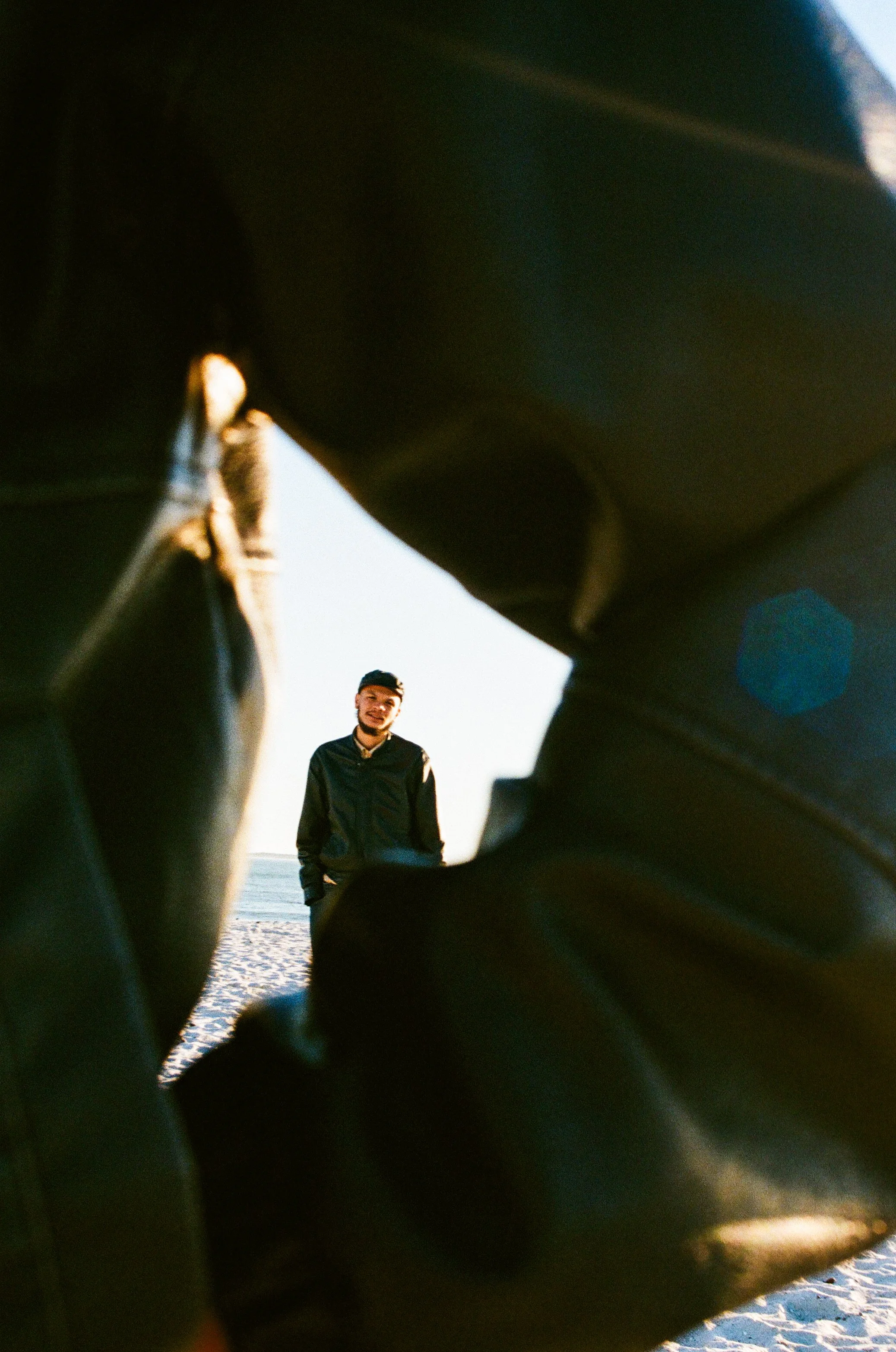 Jarry Pitboi standing on a sandy beach, seen through the gap made between the arm in the foreground, with the ocean and sky in the background.