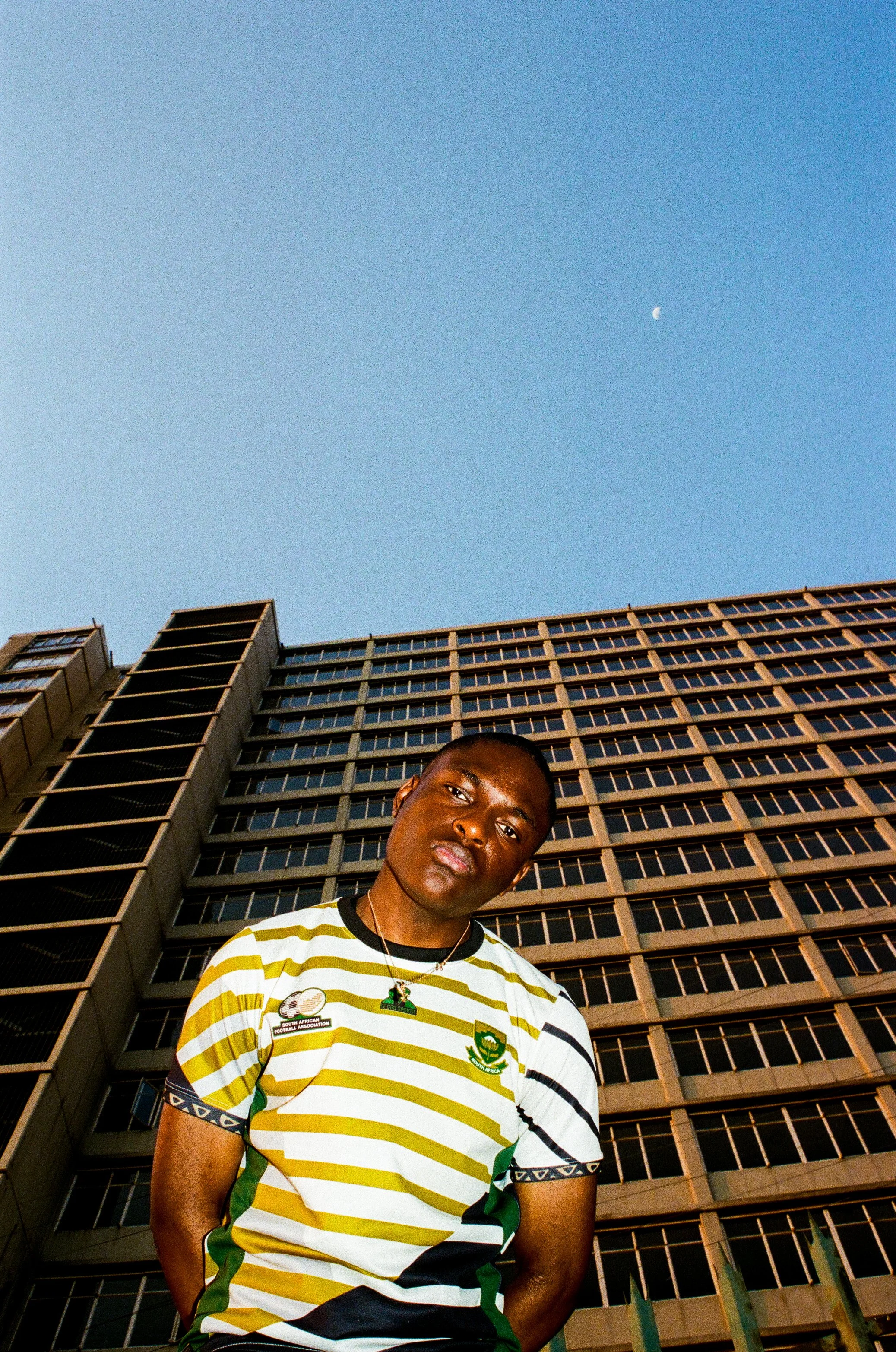 Joshua Futura standing in front of a tall building during the day, clear blue sky with a visible moon in the background.
