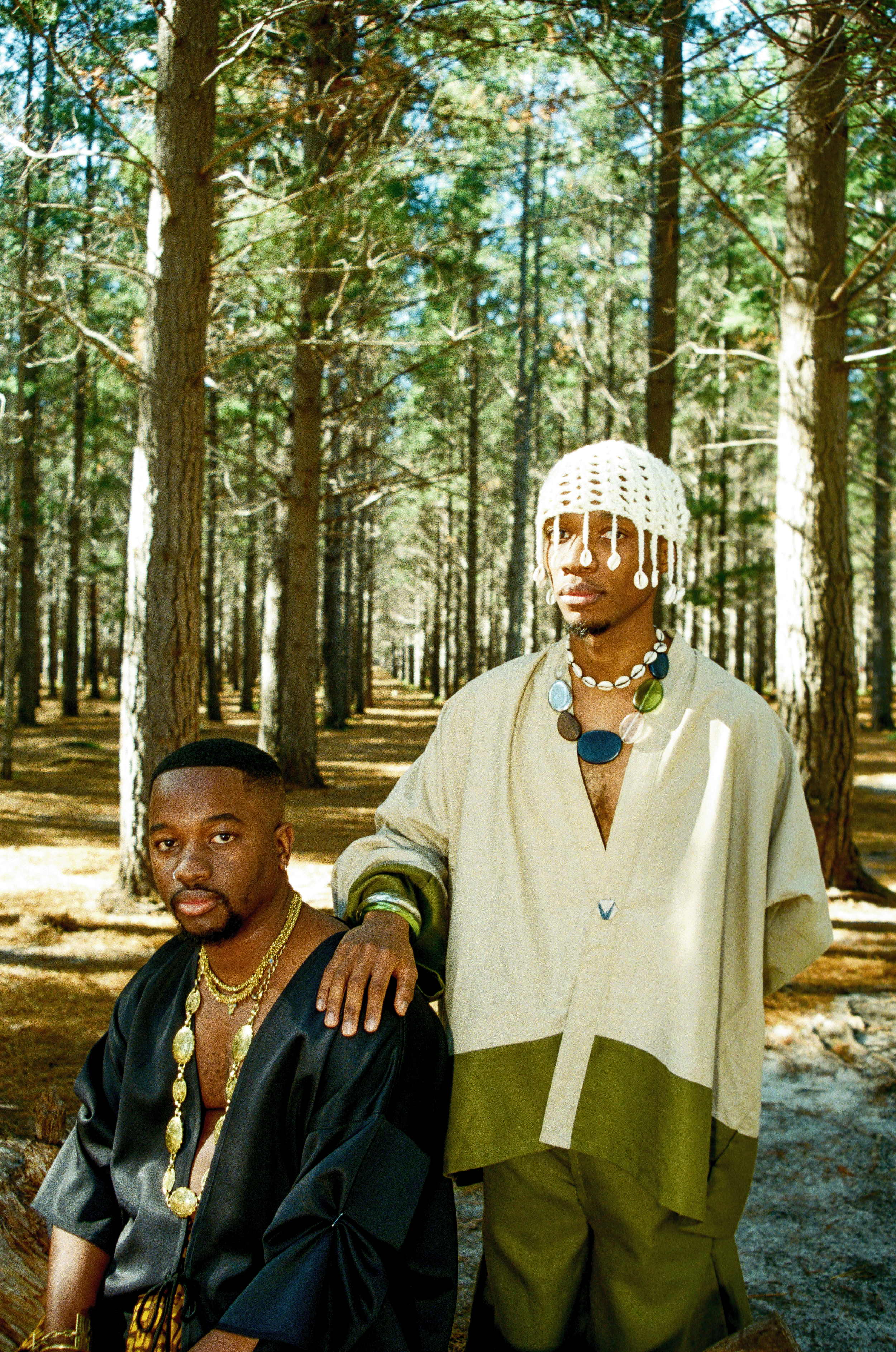 Owethu and Zwide Ndwandwe  standing in a forest with tall trees. One seated, wearing a black satin shirt and gold jewelry. The other is standing, wearing a cream and green outfit with large necklace and a knitted white hat with hanging decorations.