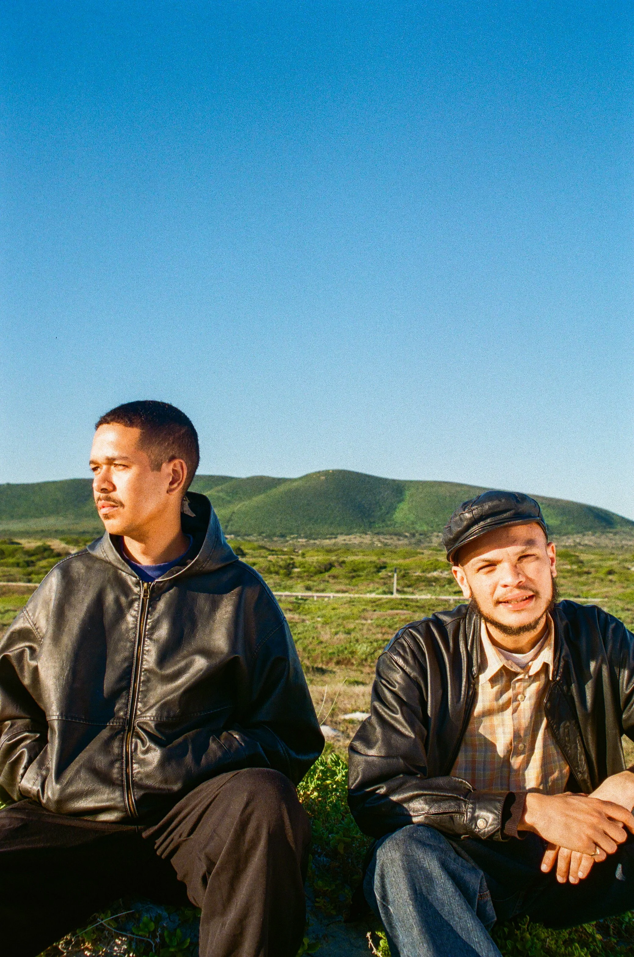 Jarry Pitboi and Permi Kwaali sitting outdoors on grassy ground with rolling green hills in the background under a clear blue sky. They are wearing black leather jackets.
