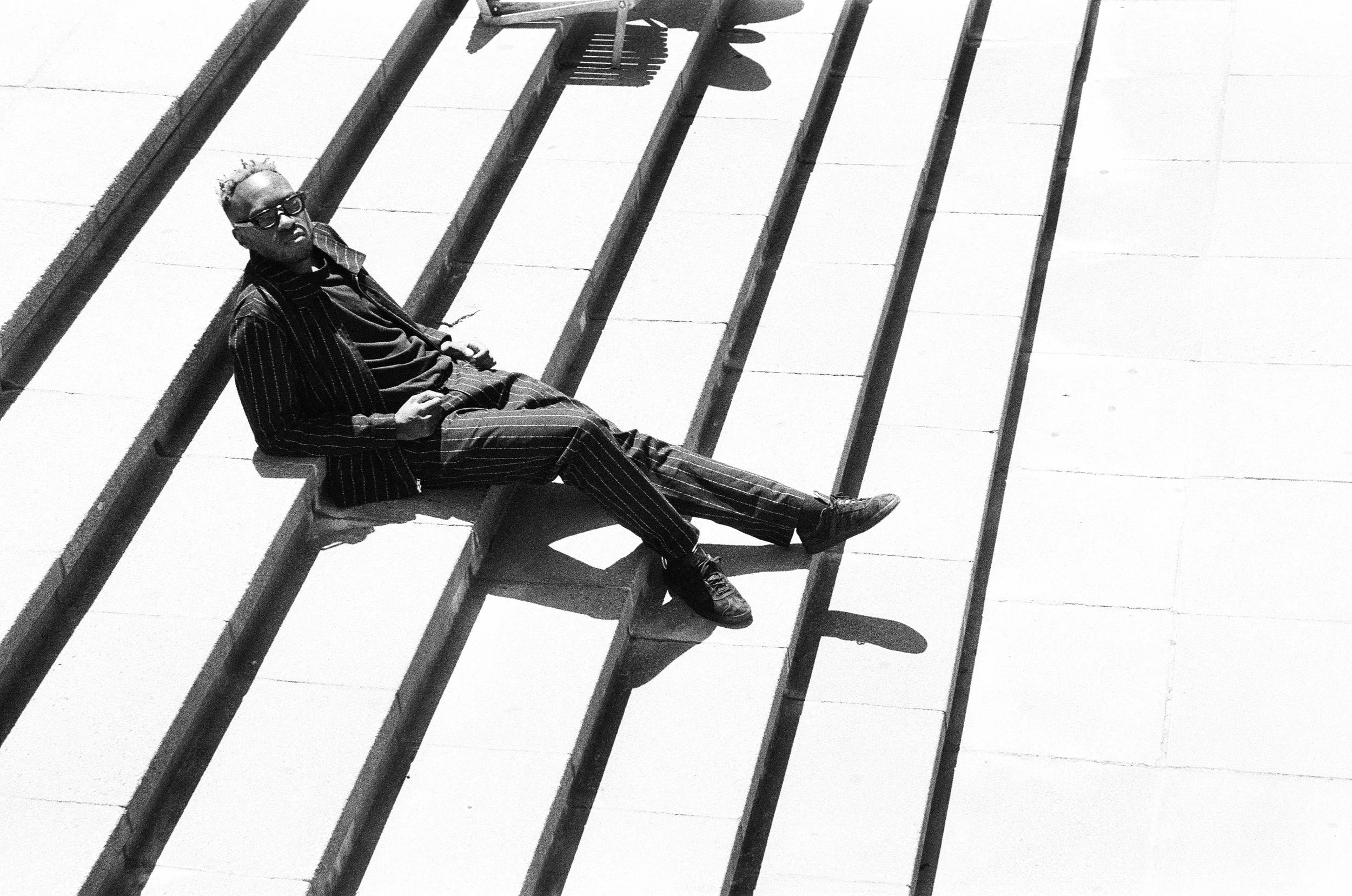 Themba Hadebe in a striped suit and dark sunglasses is lying on a bench on an outdoor staircase, with his legs stretched out and leaning back on the bench, casting a shadow on the steps.