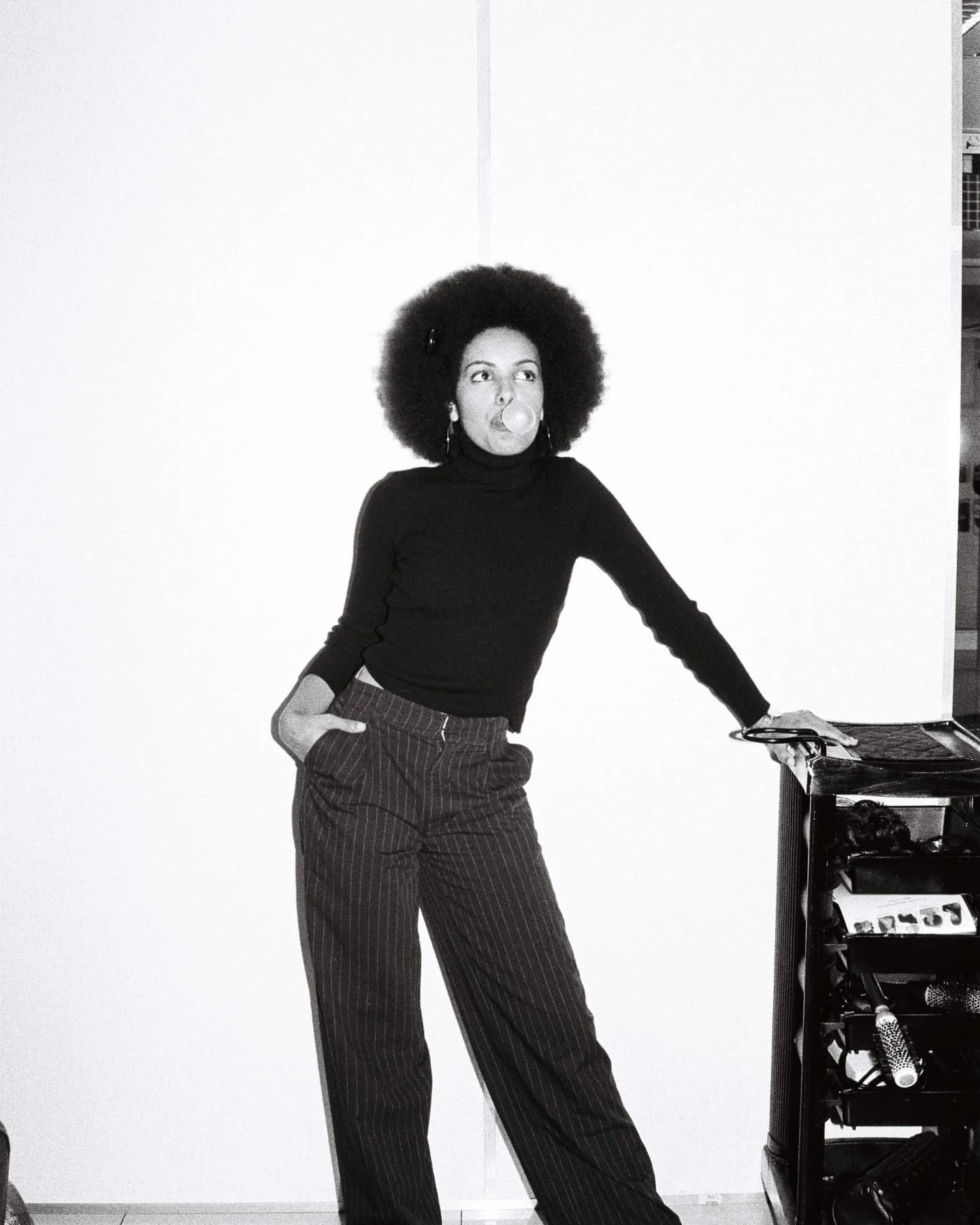 Mila Smith, with an afro hairstyle blowing bubble gum, wearing a black turtleneck and striped pants, leaning on a piano against a plain white wall.