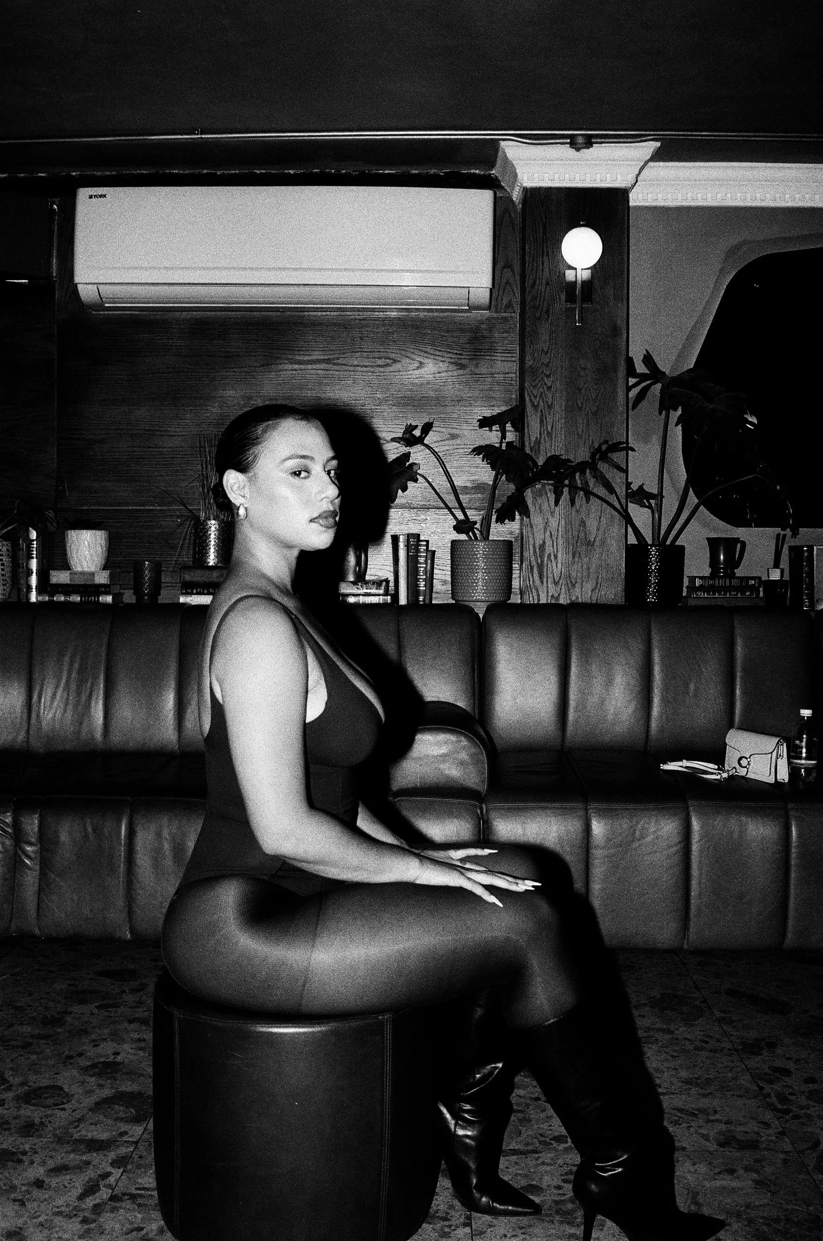 Black and white photo of a Kimberleigh Venty sitting on an ottoman in front of a leather couch, with books and plants behind her, in a room with wooden walls and framed artwork.
