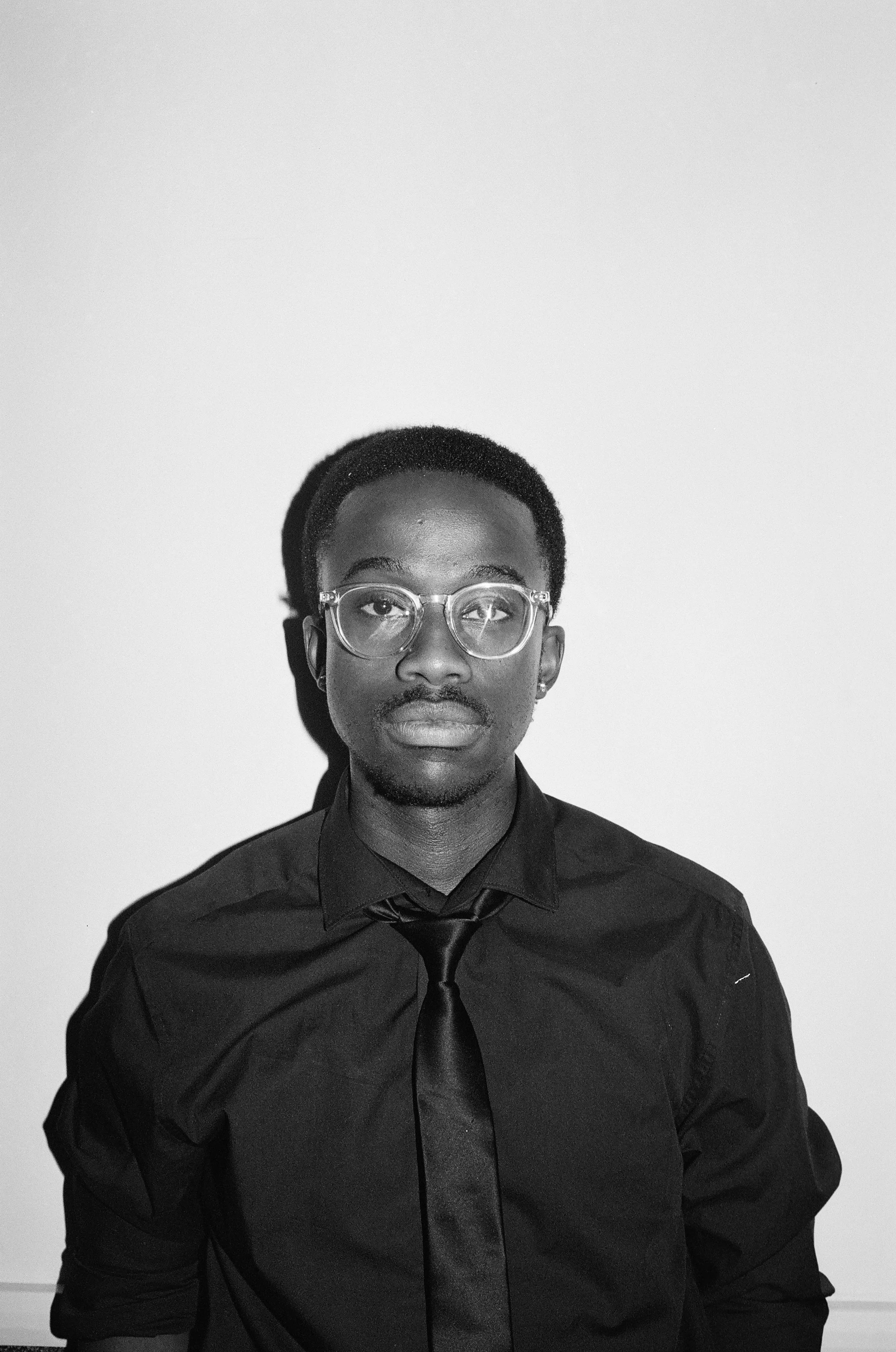 Black and white portrait of Vuyo Polson with glasses, wearing a dark shirt and tie, looking directly at the camera against a plain background.