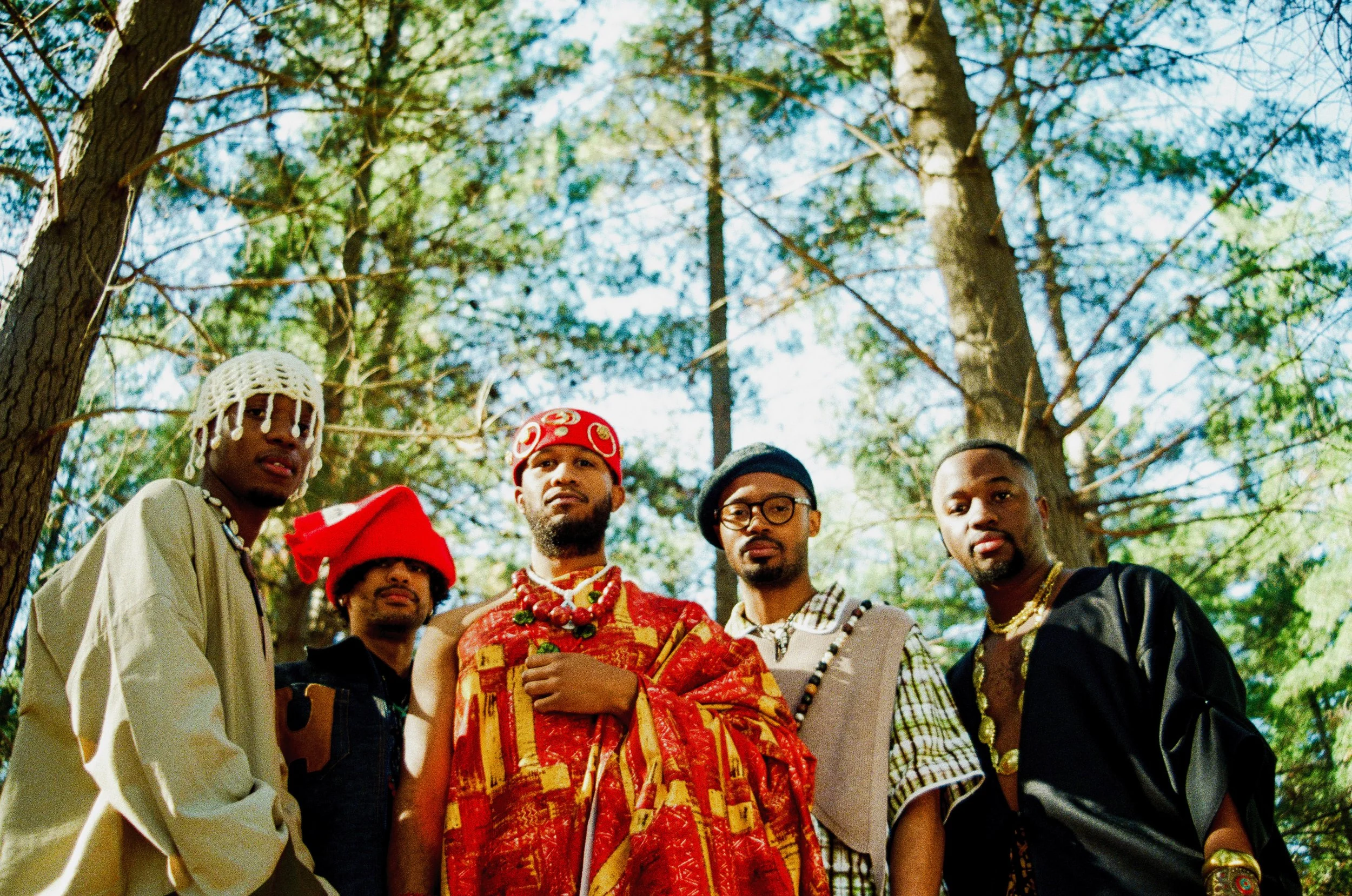 Kujenga standing outdoors under trees, wearing colorful and traditional-inspired clothing and accessories.