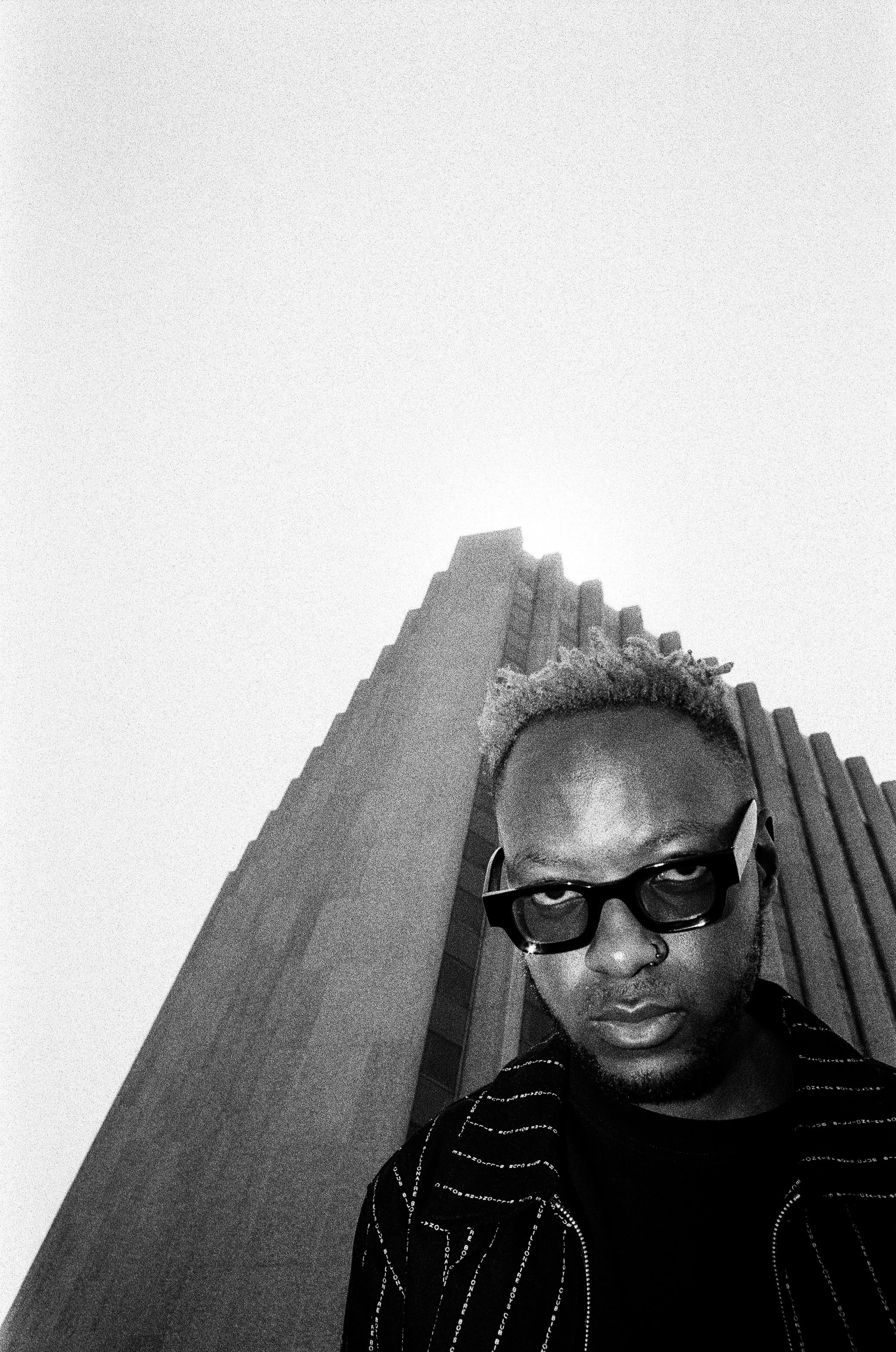 Black and white photo of a man, Themba Hadebe, with light-colored hair, glasses, and a nose ring, standing in front of a tall, modern building with vertical architectural elements.
