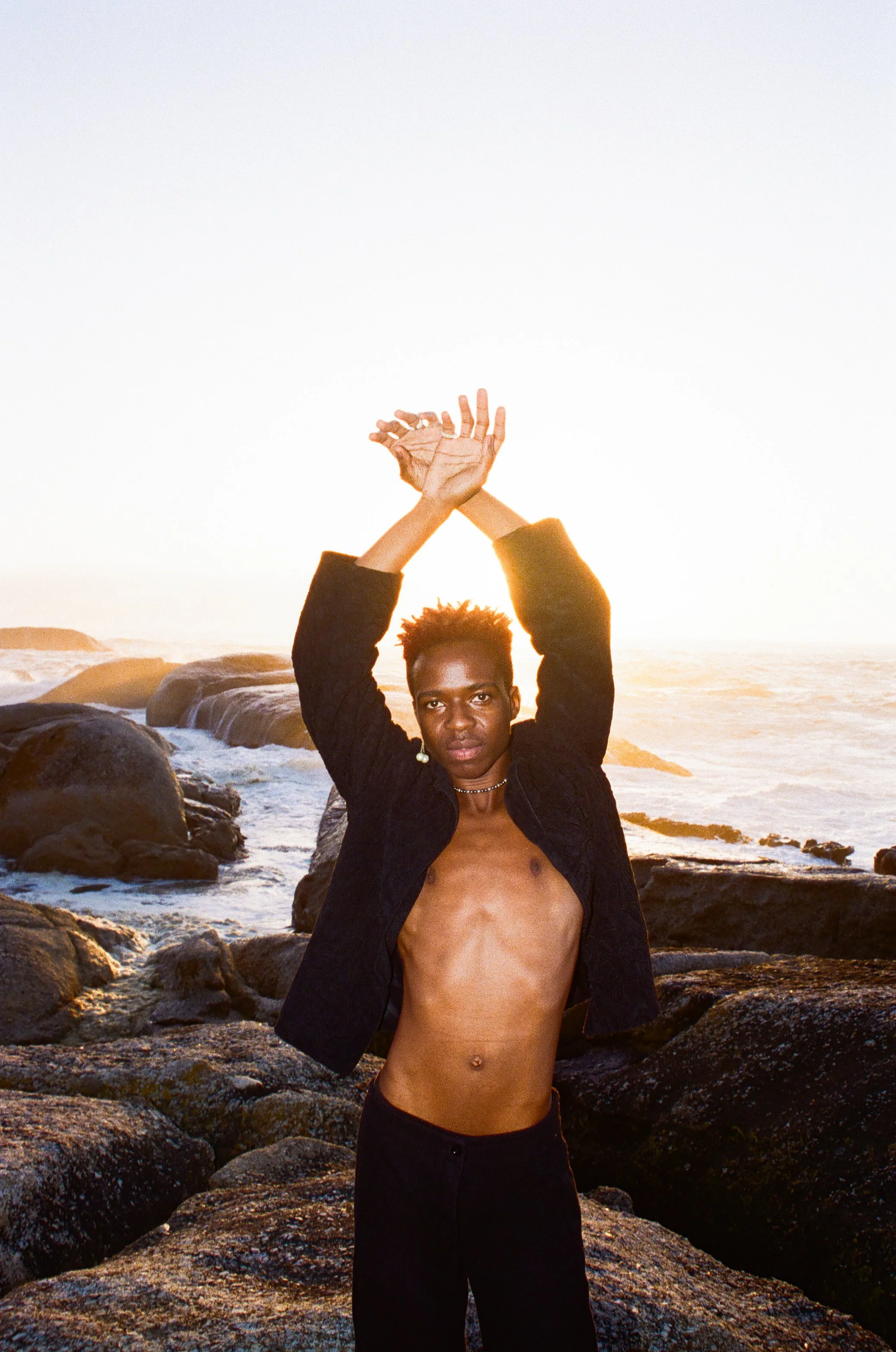Dnemas standing on rocks near the ocean at sunset with hands raised above their head.