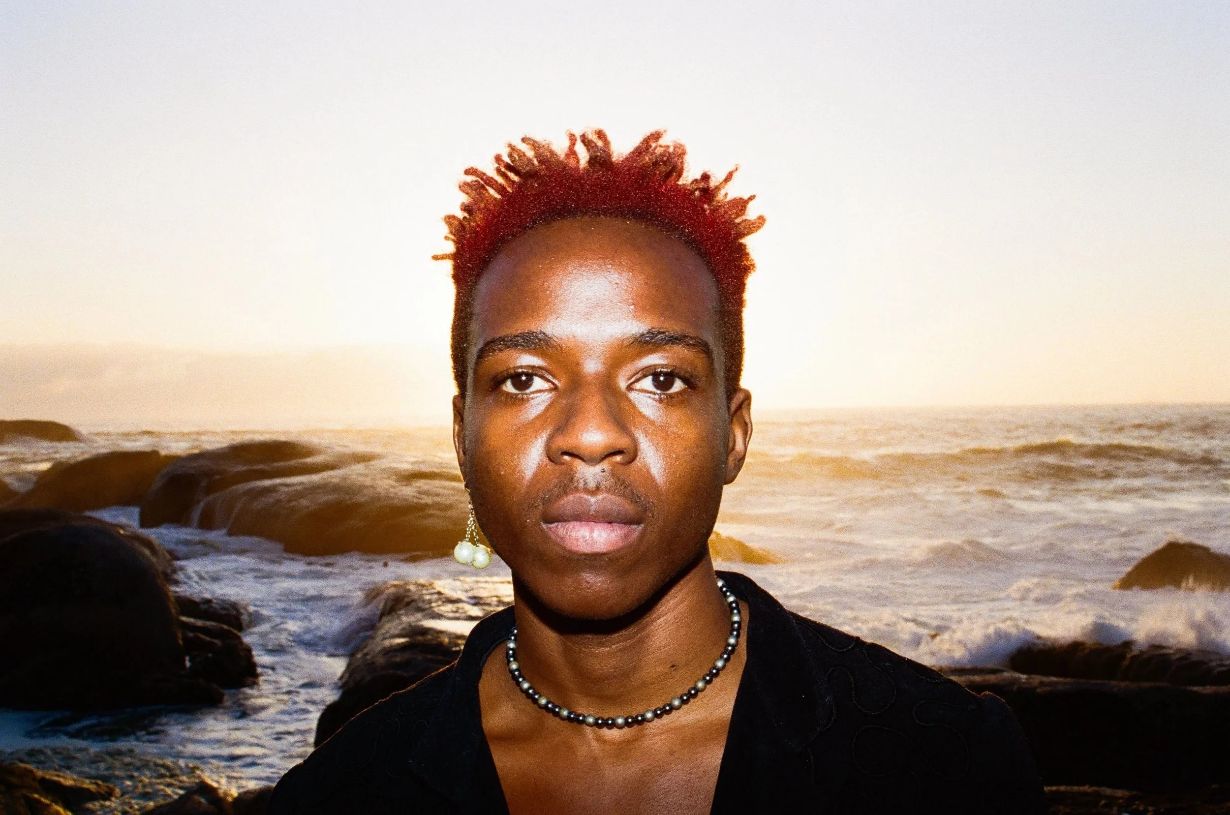 Dnemas with short, bright red hair standing on a rocky beach during sunset, wearing a black top, pearl earrings, and a pearl necklace.