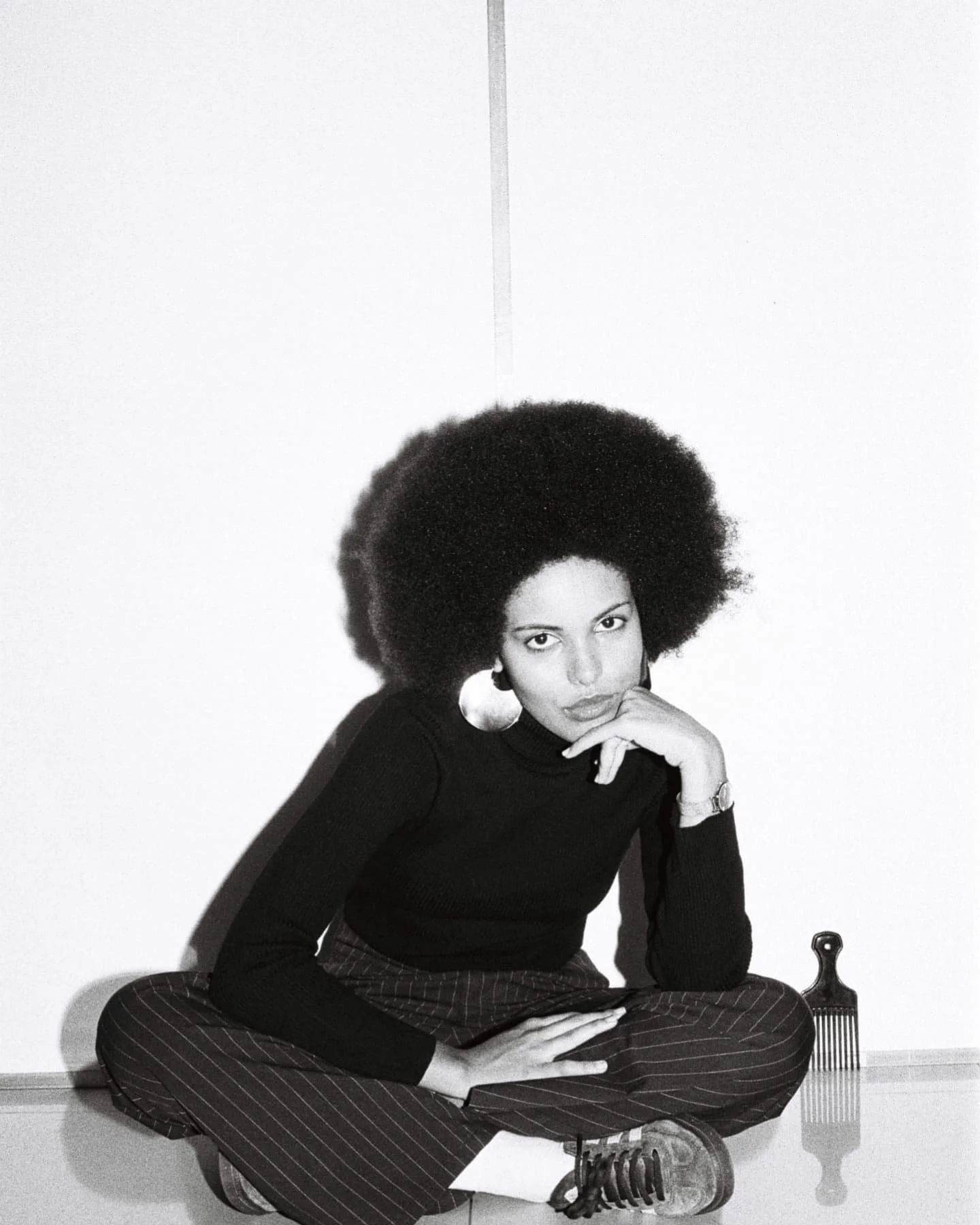 A Black and white photo of a woman with large afro hairstyle sitting on the floor in front of a plain wall. She is wearing a long-sleeved black top, pinstriped pants, and sneakers. She has large hoop earrings, a watch, and a ring, with one hand resting on her chin and the other on her knee. There is a hair comb on the floor beside her.