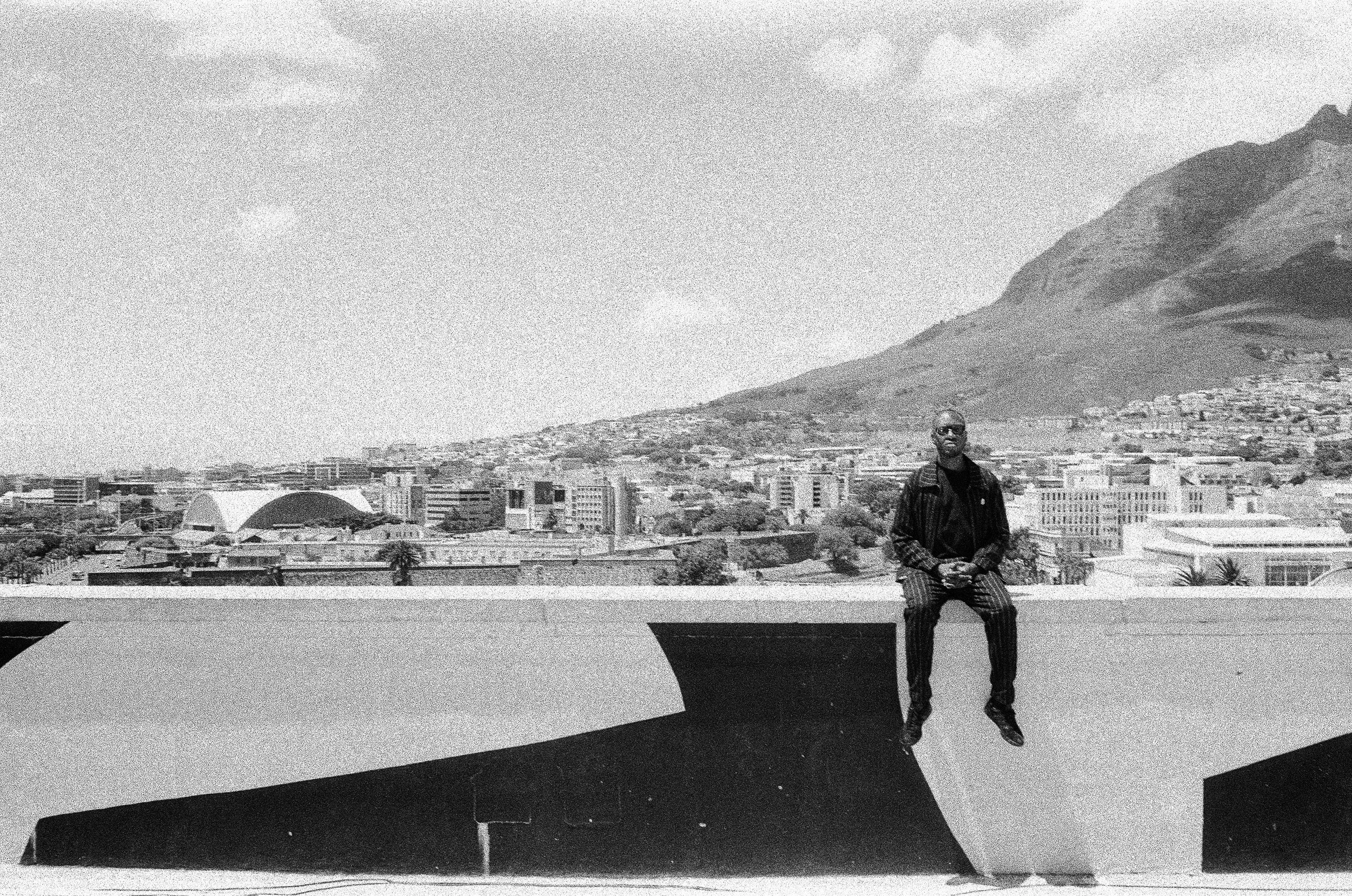 Themba Hadebe sitting on the edge of a rooftop with a cityscape and mountain in the background, in black and white.
