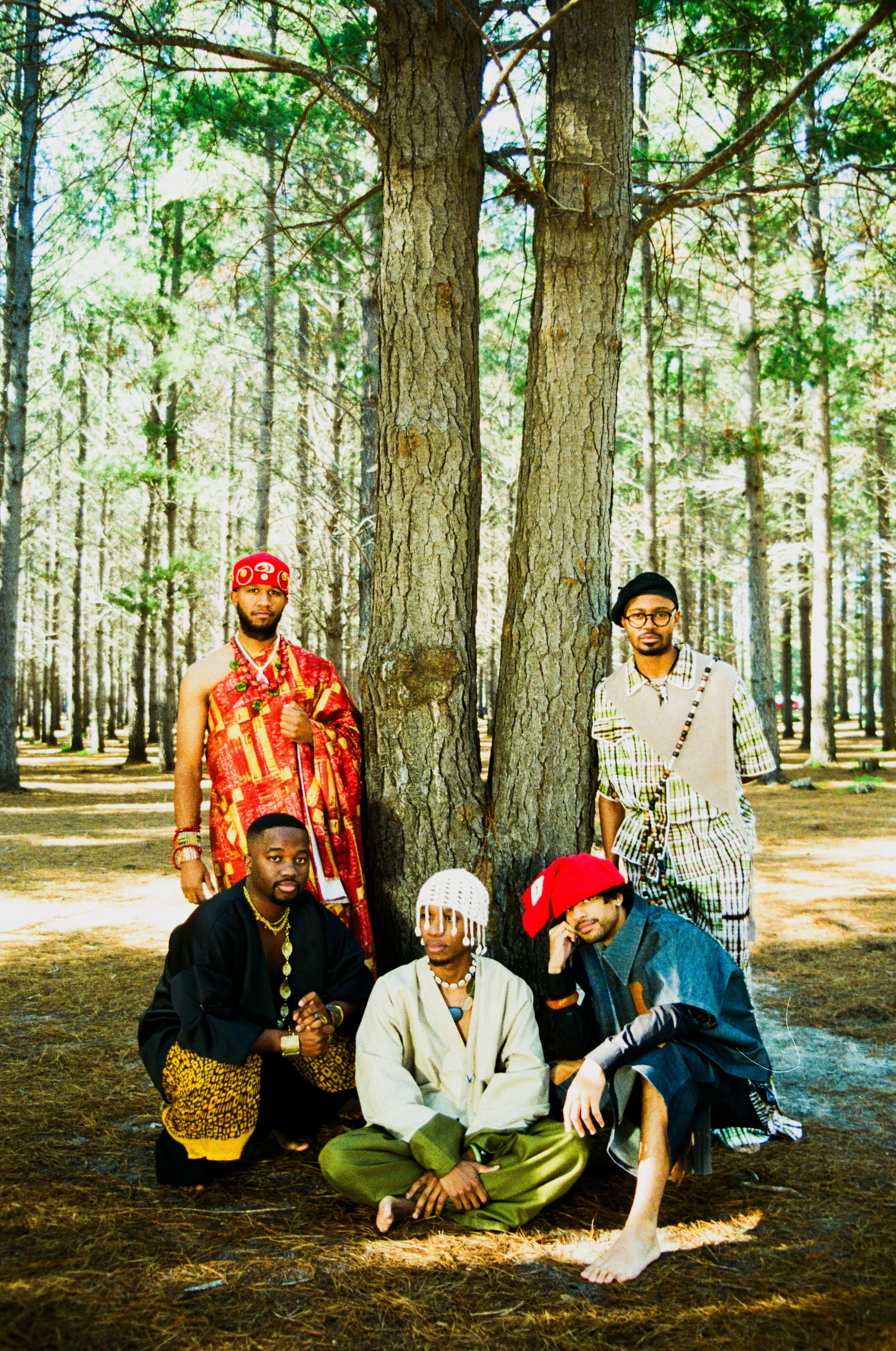 Kujenga in colorful, eclectic clothing pose in a wooded area with tall trees, some sitting and some standing.