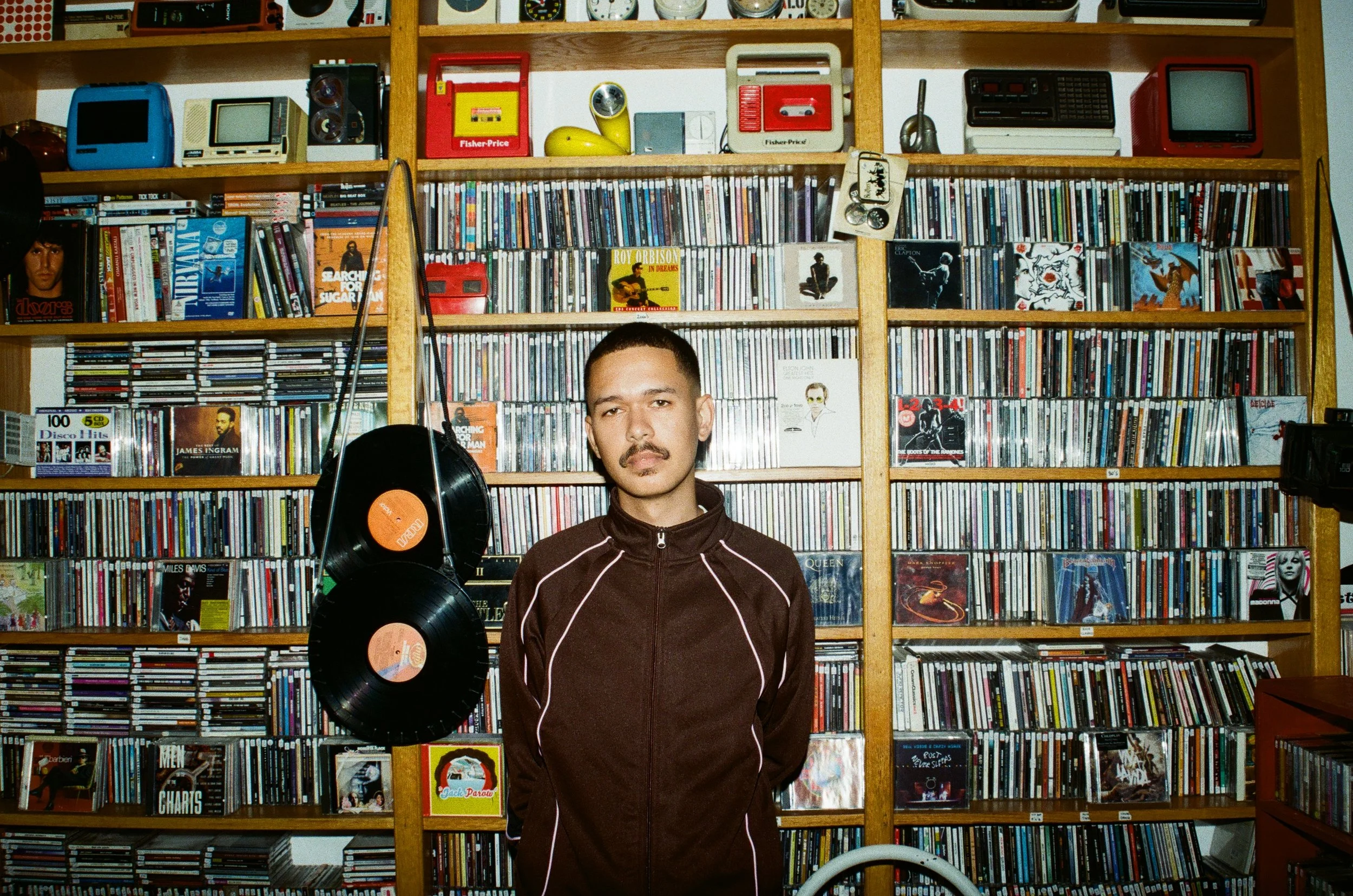 Jarry Pitboi standing in front of a large wooden bookshelf filled with CDs, records, and vintage electronics. He wears a dark zip-up jacket with white piping, has a mustache, and short hair.