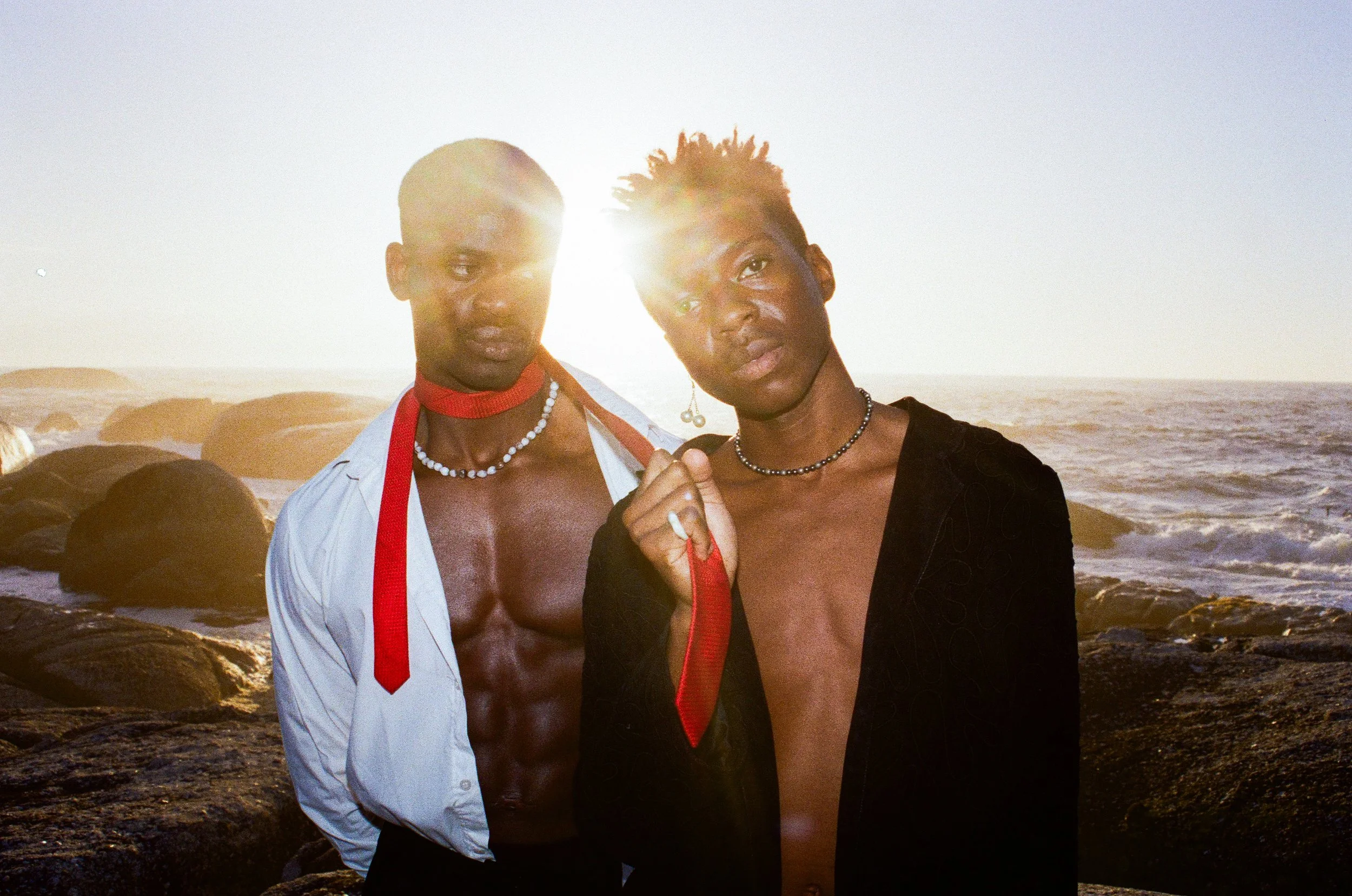 Two young Black men standing on a rocky beach at sunset, one shirtless with a red tie around his neck and the other wearing a black jacket with no shirt, holding a red tie, both with necklaces and earrings, with the sun setting behind them over the ocean.