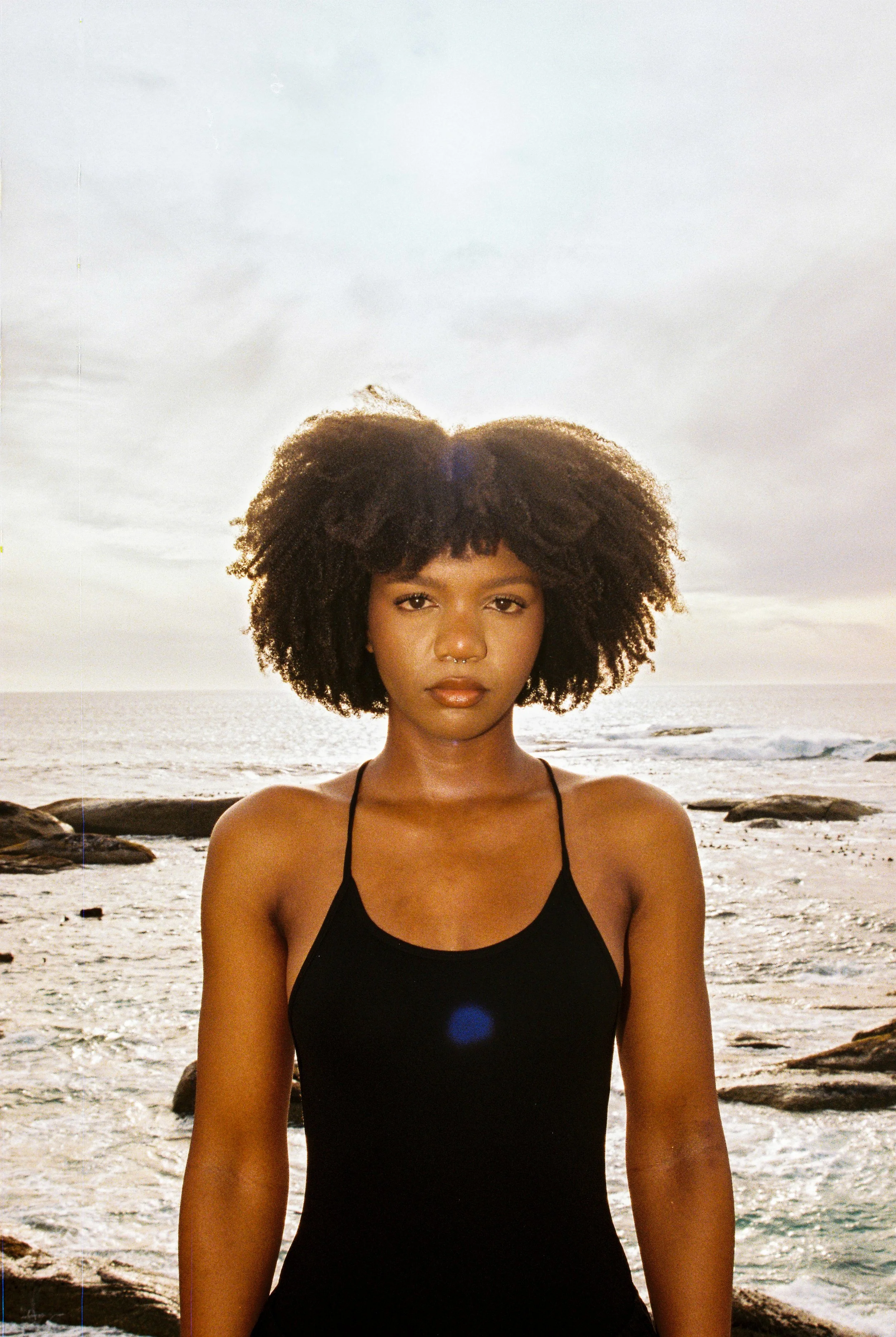 A woman with natural curly hair stands on a rocky beach during sunset, wearing a black tank top and looking directly at the camera.