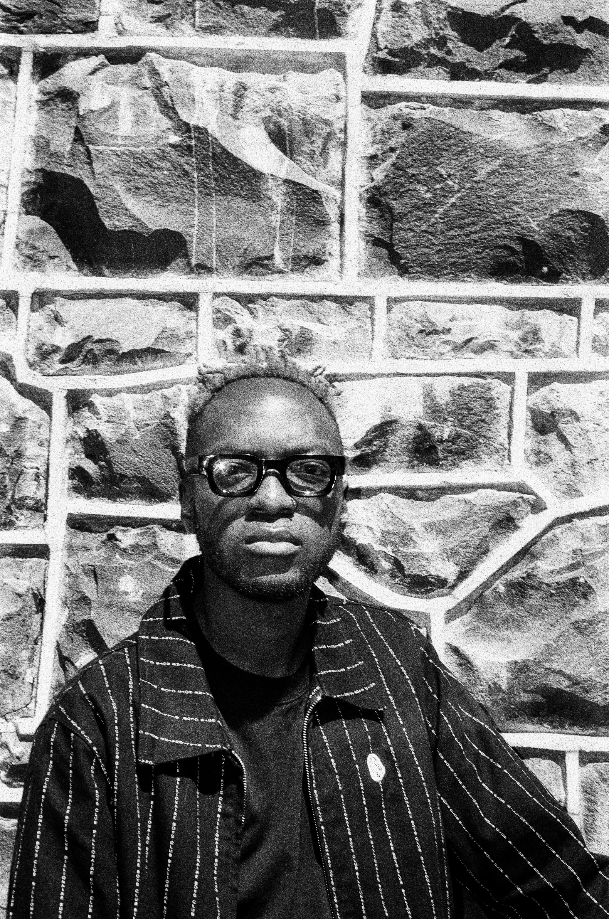 Themba Hadebe with glasses and a striped jacket standing against a textured stone wall in black and white.