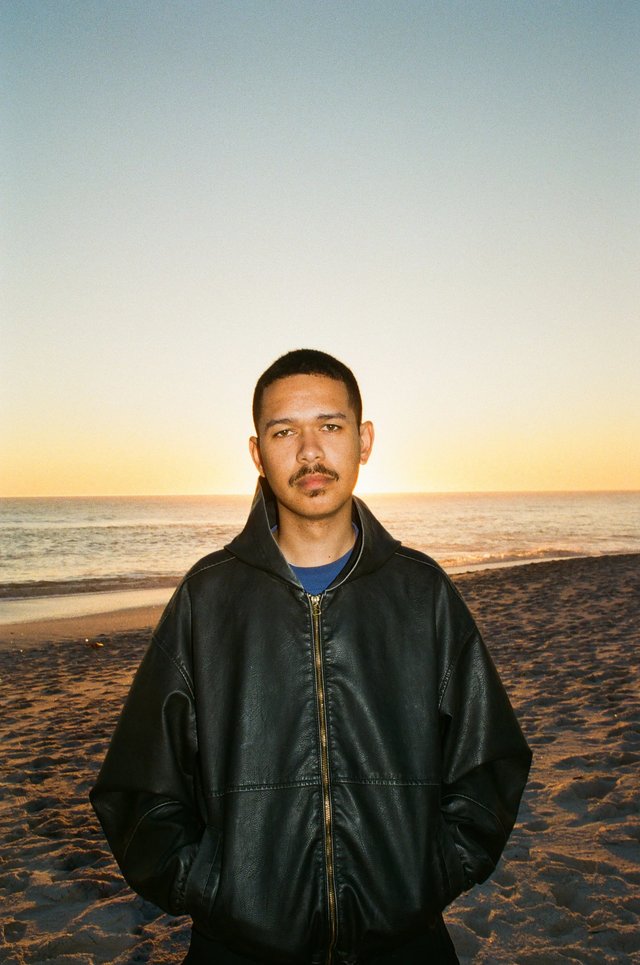 Jarry Pitboi standing on a beach at sunset, wearing a black leather jacket.