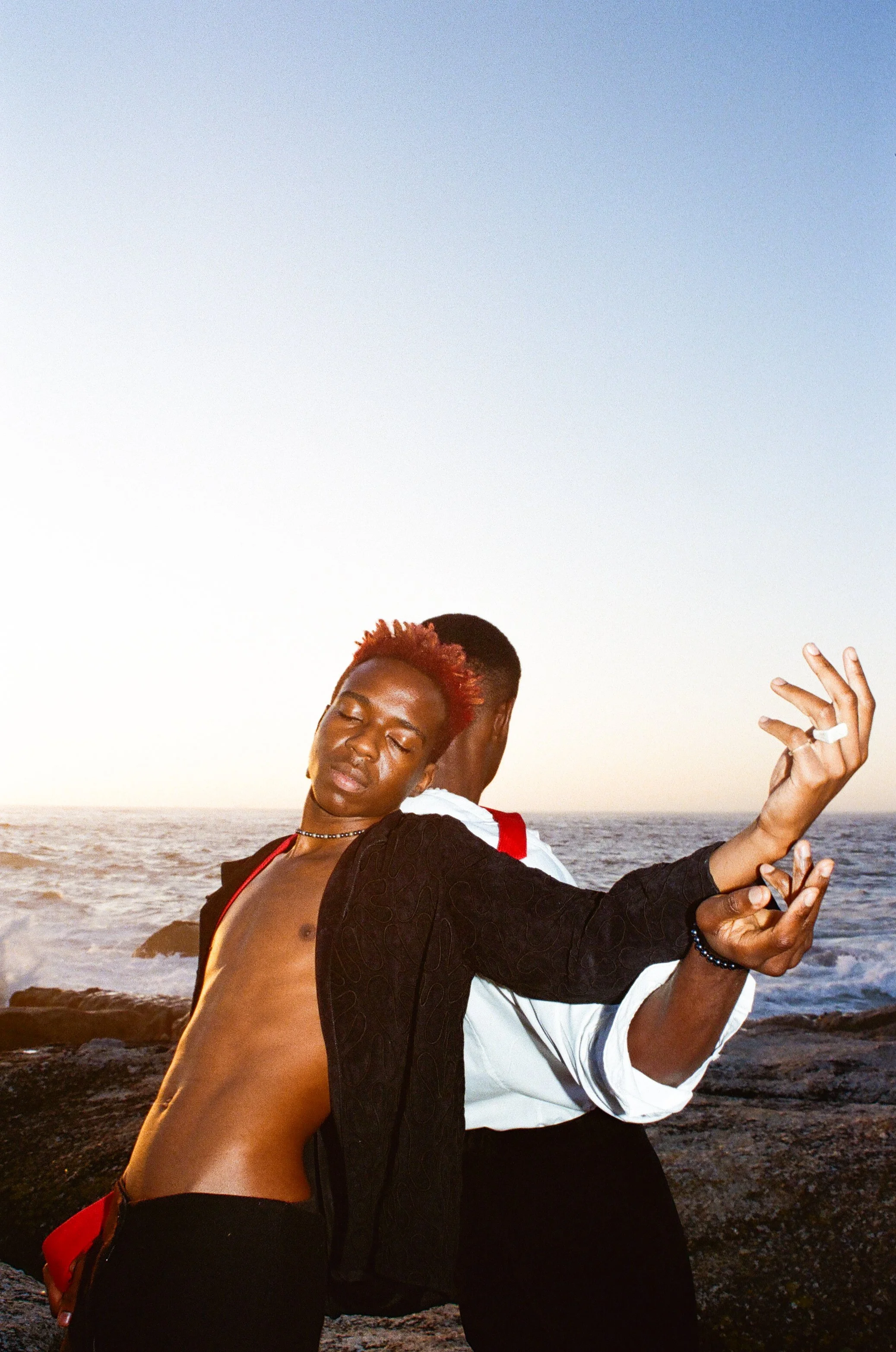 Two people dancing passionately on rocks by the ocean at sunset, with Dnemas person having red hair and the other turned away, creating a dramatic and intimate scene.