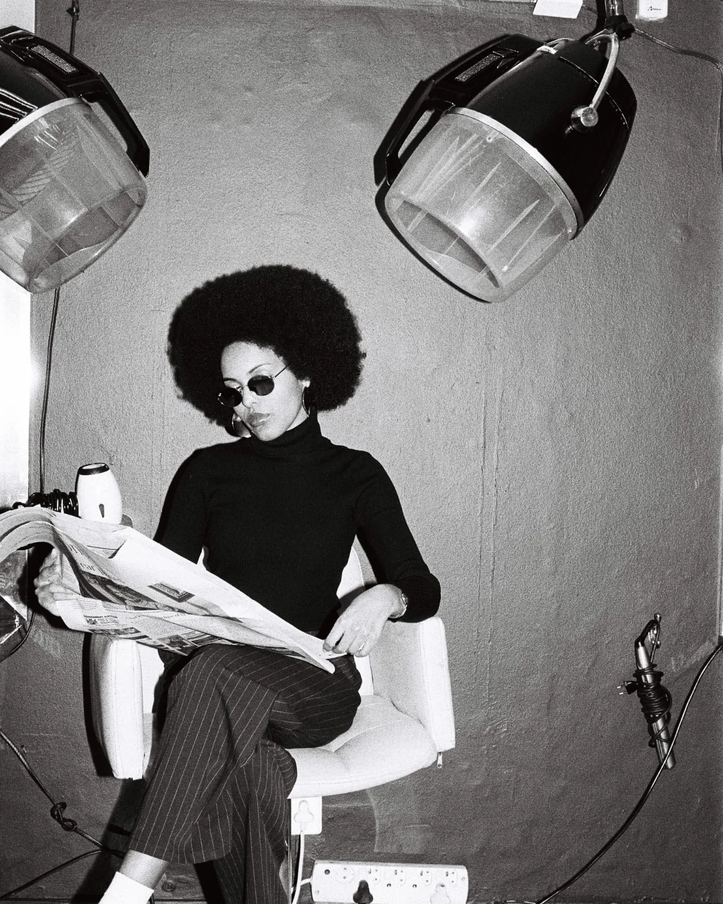 Mila Smith wearing an afro hairstyle, wearing dark sunglasses, a black turtleneck, and pinstriped pants, is sitting in a salon chair reading a newspaper. The salon has hair drying/steaming machines hanging from the ceiling.