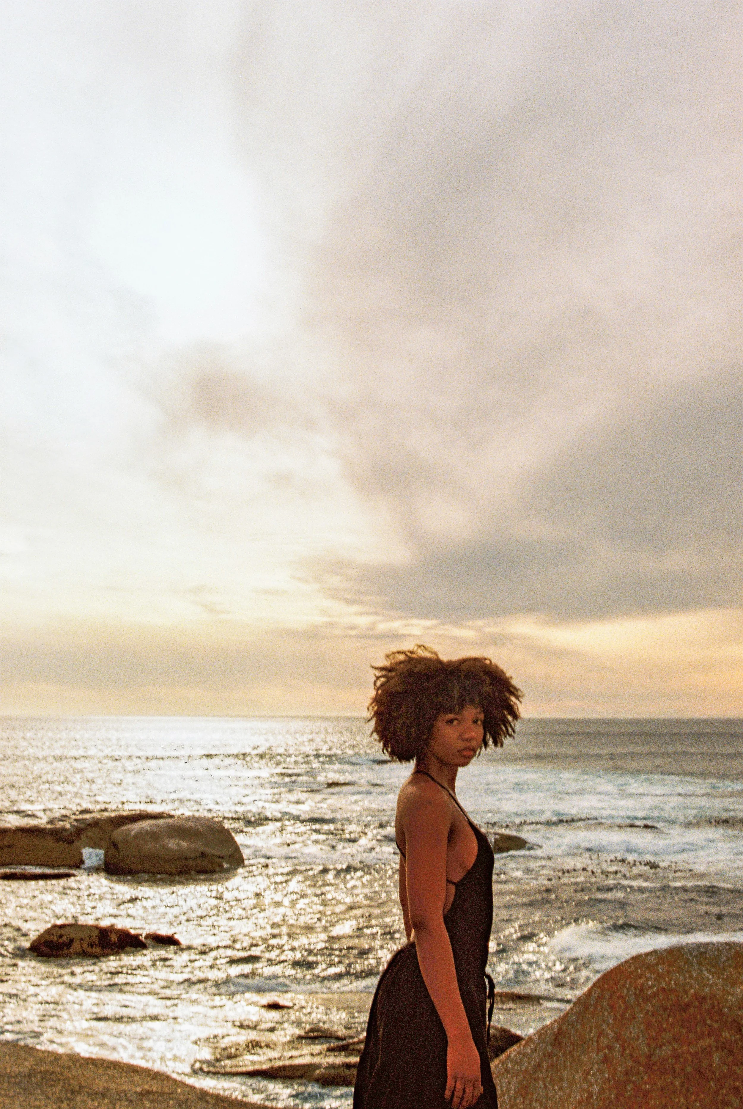 A woman with curly hair standing on the rocky shoreline during sunset, ocean waves behind her.