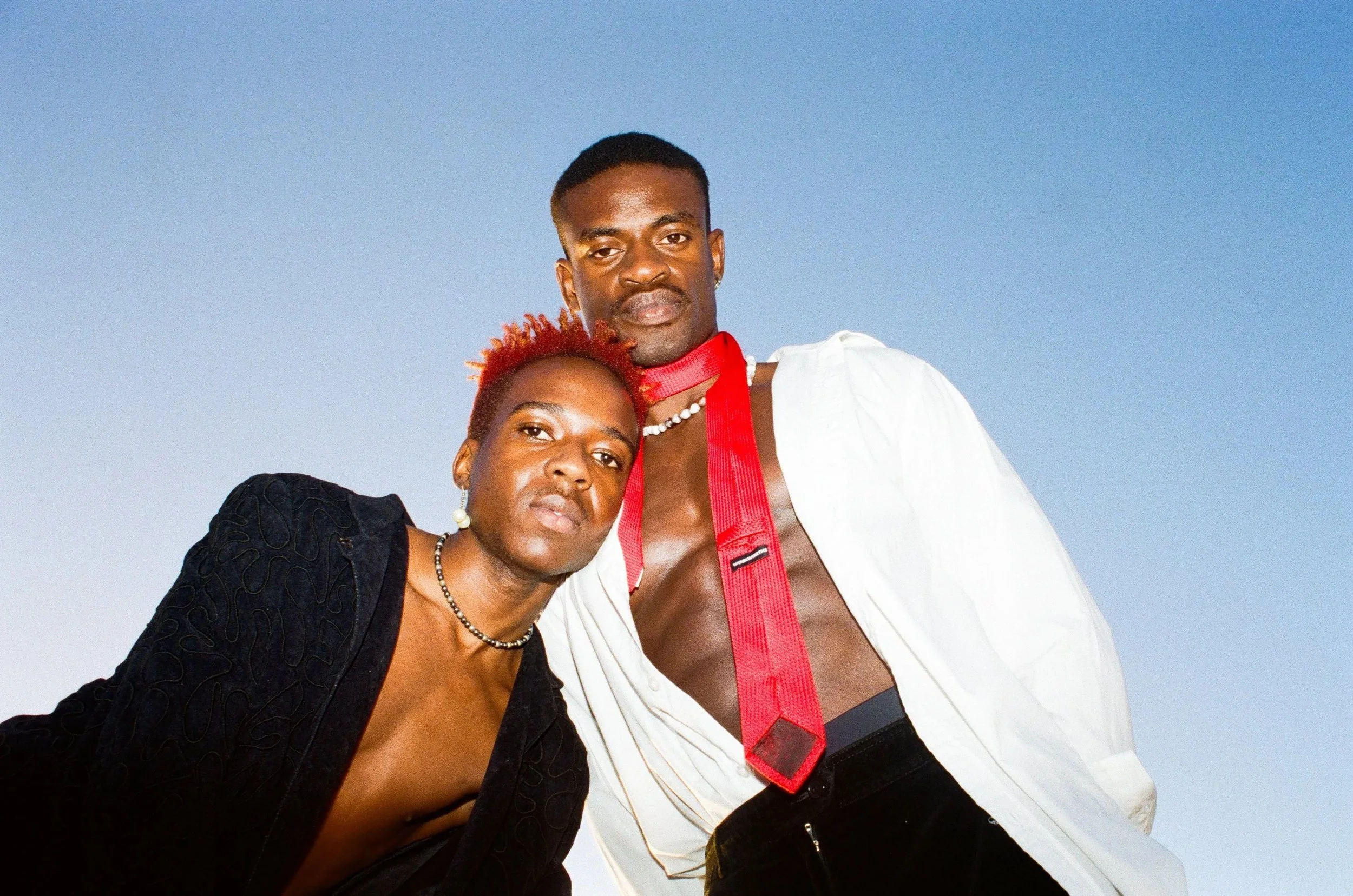 Dnemas and a friend posing against a clear blue sky, one with short bright red hair wearing a black jacket, the other with short black hair in a white open shirt with a red tie hanging loosely, both looking at the camera.