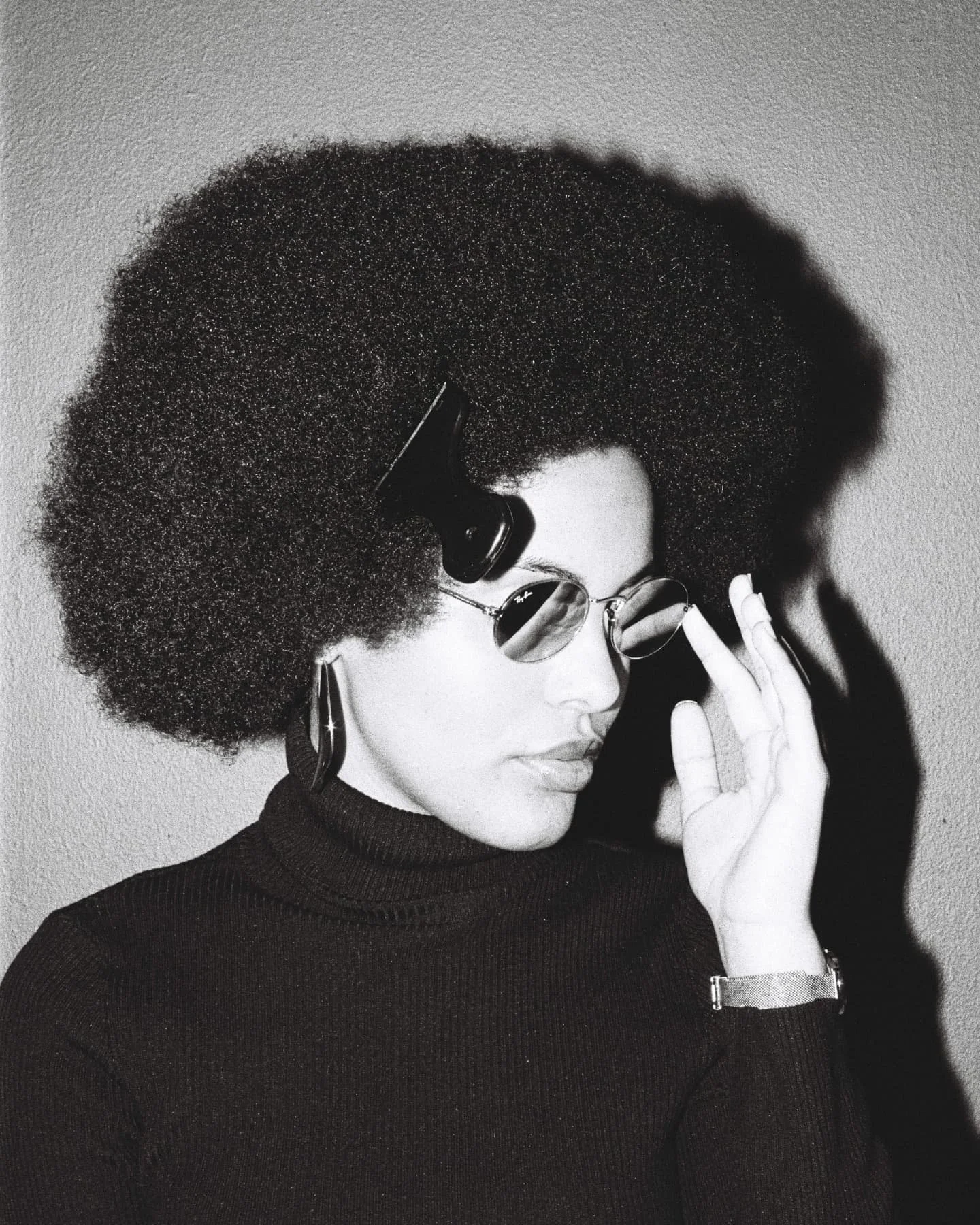 Black and white photo of Mila Smith wearing round sunglasses, large afro hairstyle, earrings, and a turtleneck sweater, touching her sunglasses.