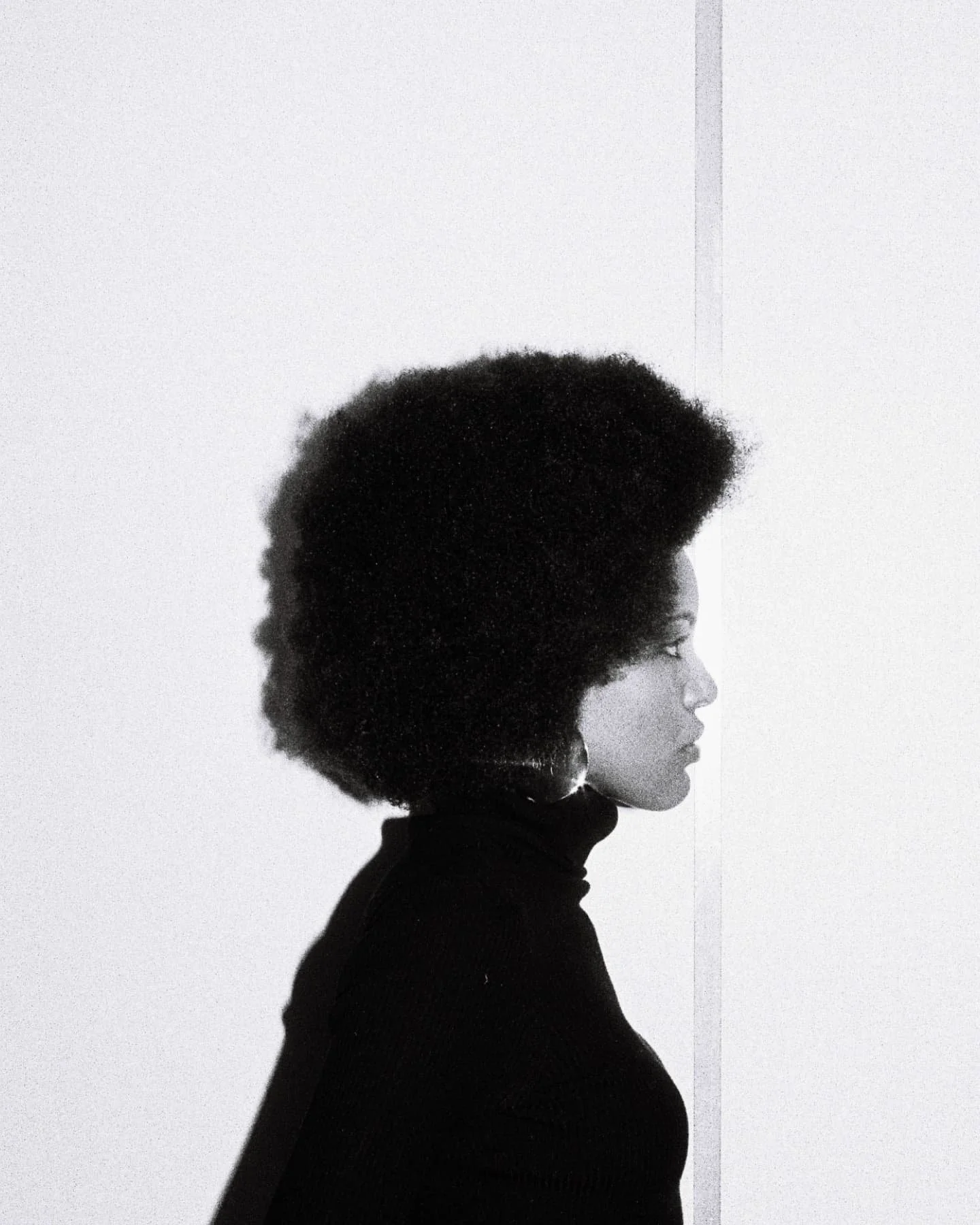 Side profile of Mila SMith with natural afro hairstyle wearing a black turtleneck against a plain white background.