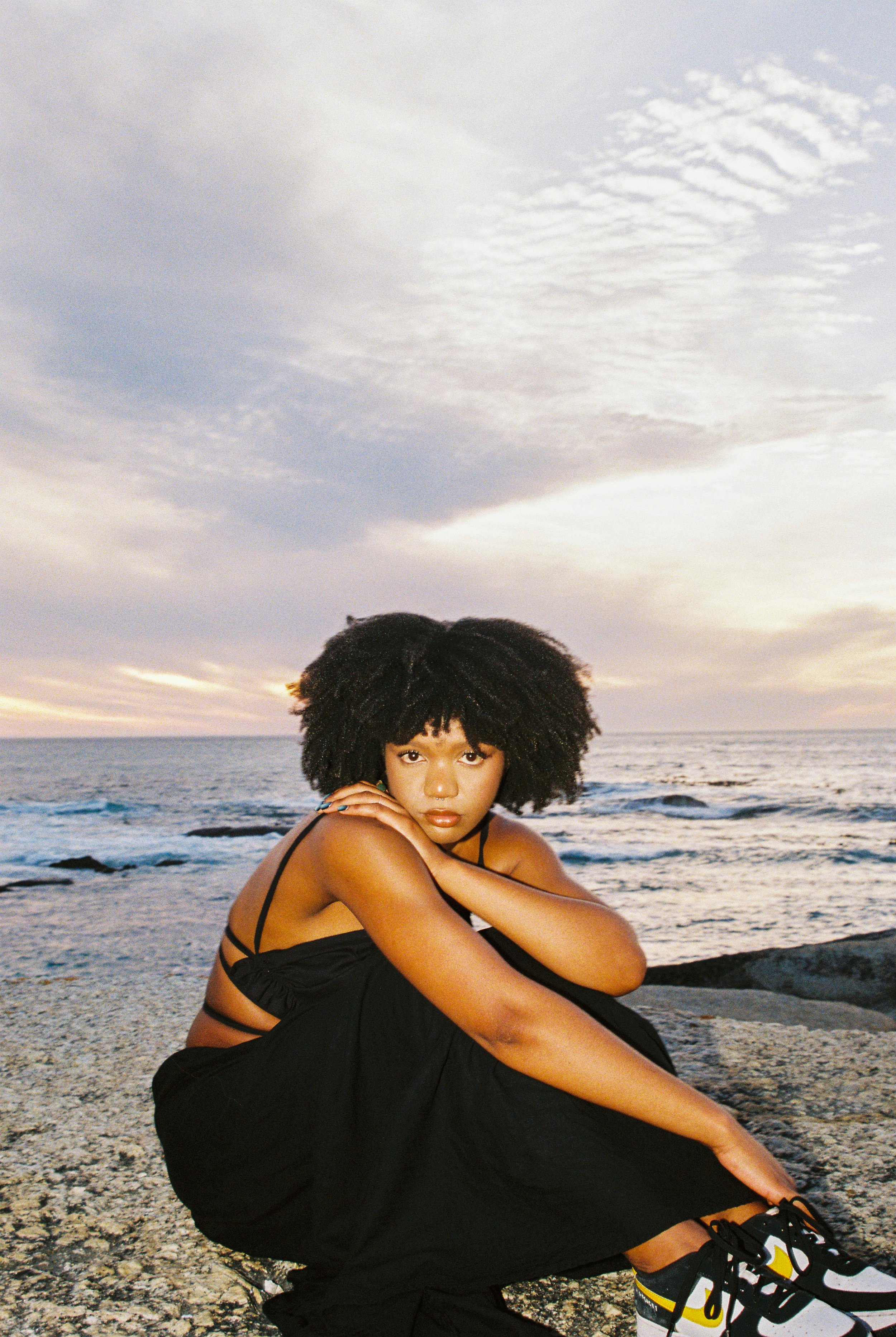 A young woman with curly black hair wearing a black dress and sneakers, sitting on rocks by the ocean during sunset, looking directly at the camera.