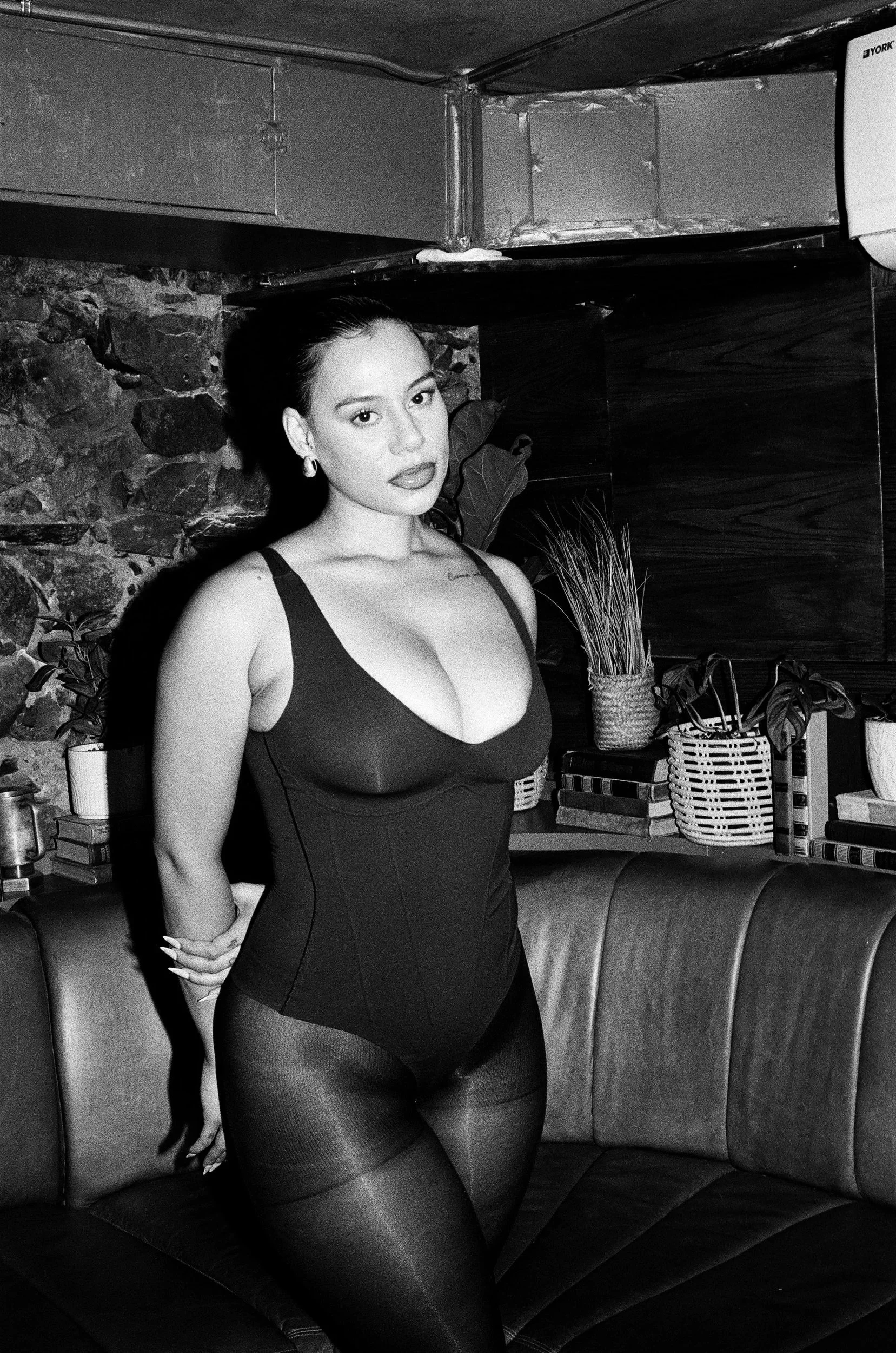 Kimberleigh Venty posing in a black outfit, in a room with a stone wall and plants, in black and white.