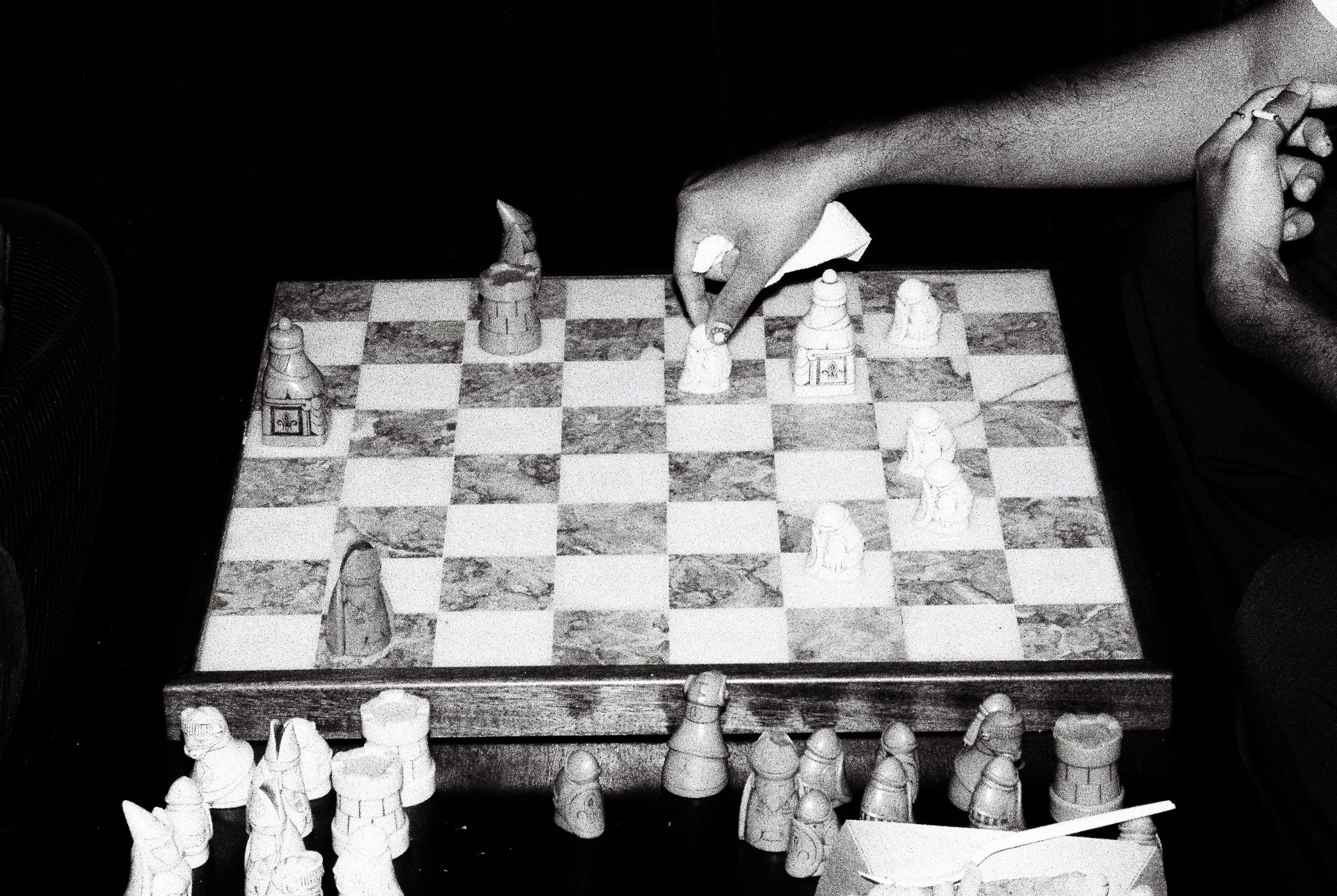 A person is playing chess on a chessboard, with several chess pieces arranged on the board and on the table around it. The immediate view shows the person's arms and hands, with one hand holding a chess piece over the board.