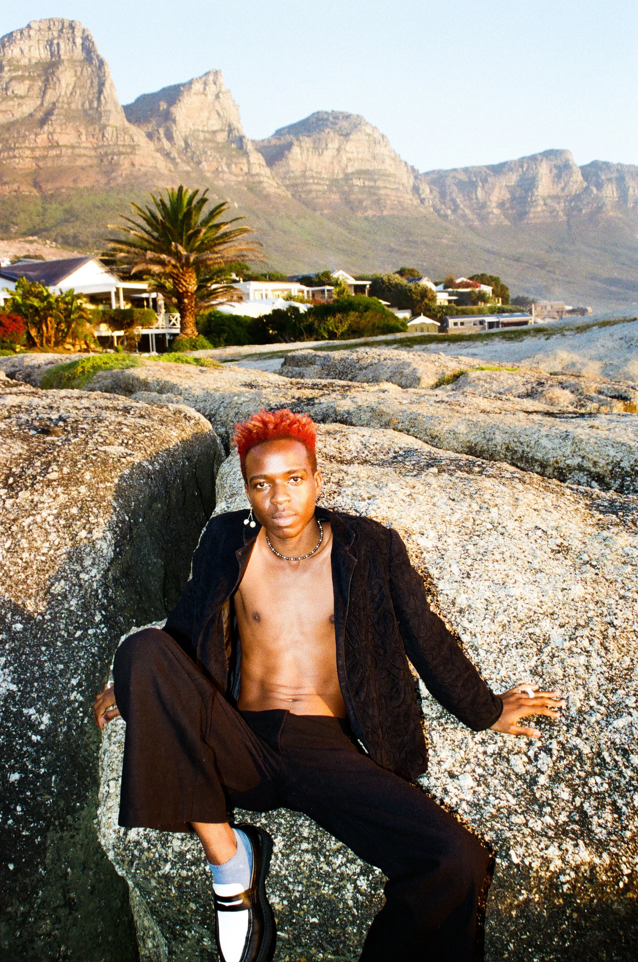 Dnemas with short, bright red hair sitting on rocks in a coastal area with mountains, houses, and greenery in the background.