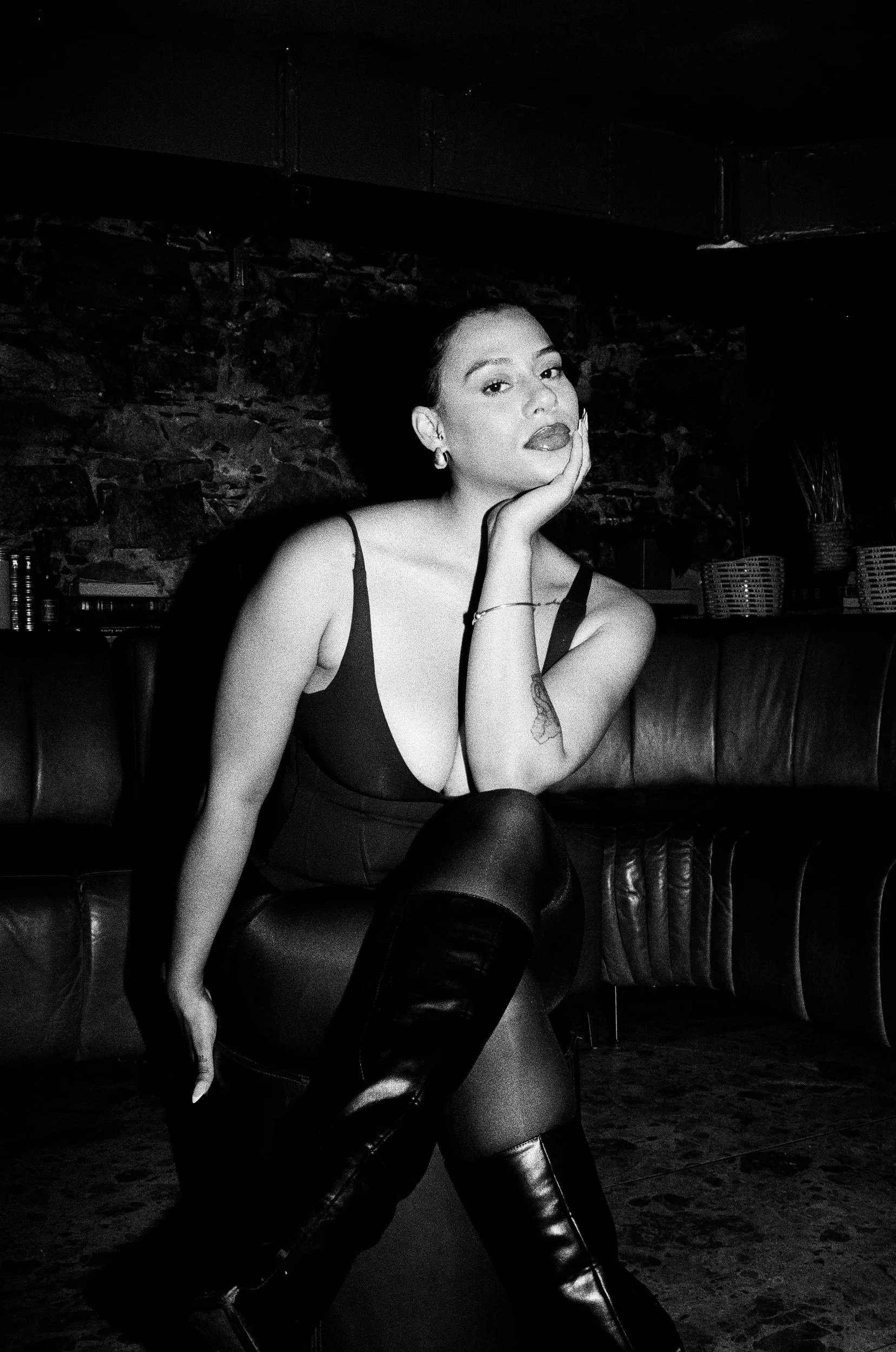 Black and white photo of Kimberleigh Venty sitting on a leather sofa, resting her head on her left hand, wearing a low-cut top and knee-high leather boots.