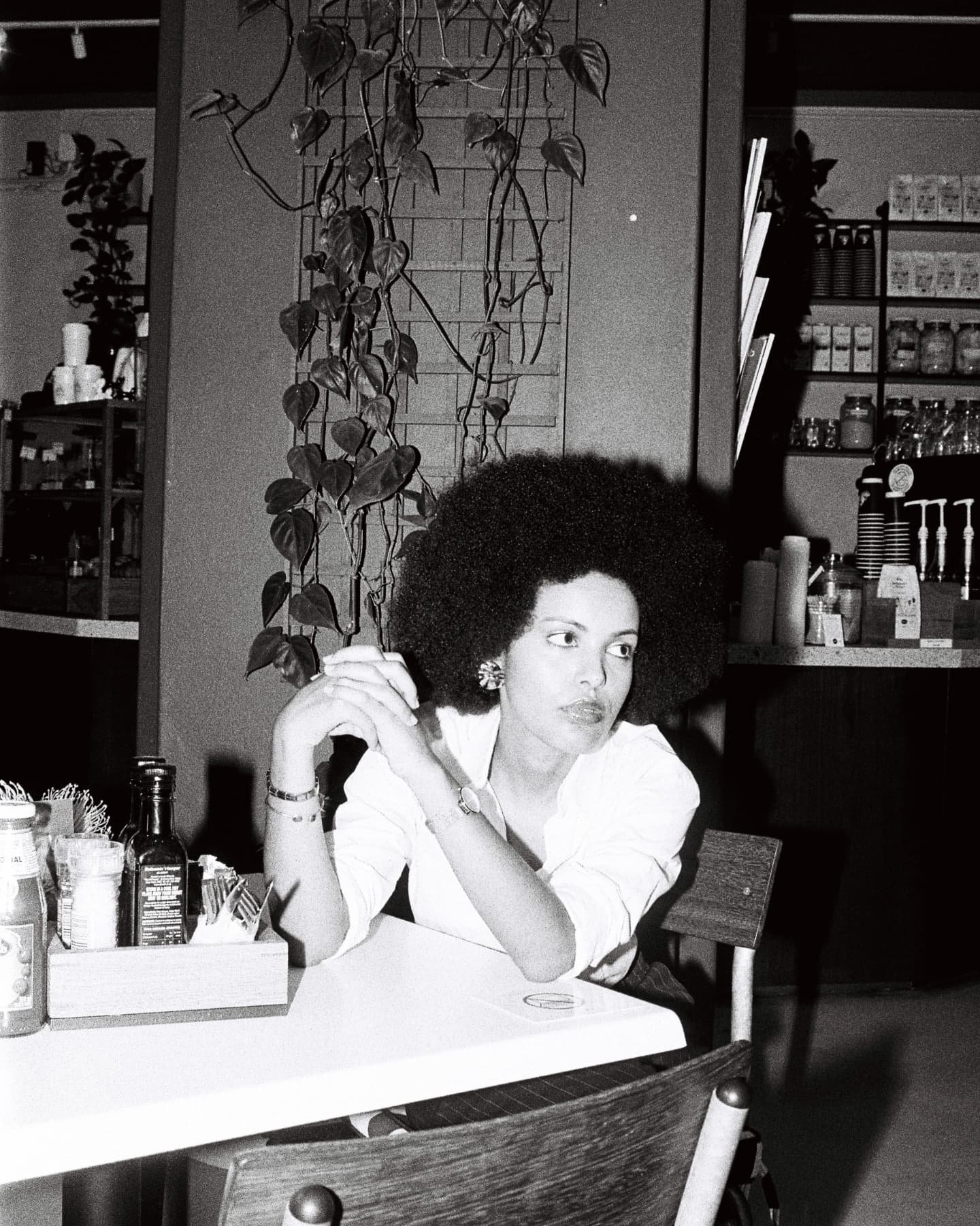 Mila Smith with a large afro hairstyle sitting at a table in a cafe, wearing earrings and a white shirt, resting her arms on the table, looking to the side.