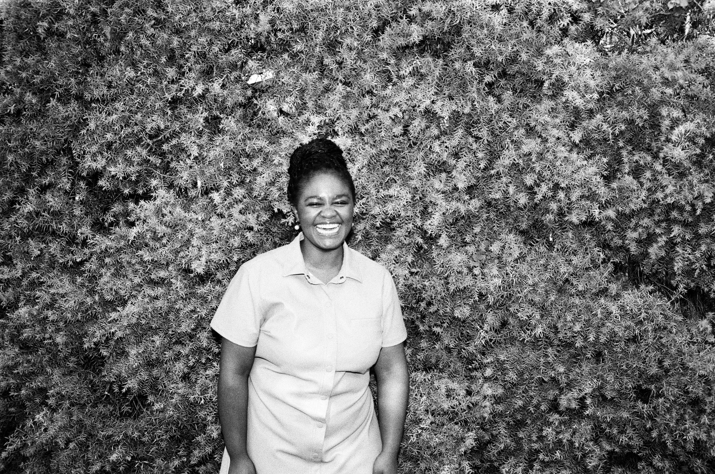 A smiling woman, Zizipho Bam, hair standing in front of dense, bushy foliage. Her hair braided.