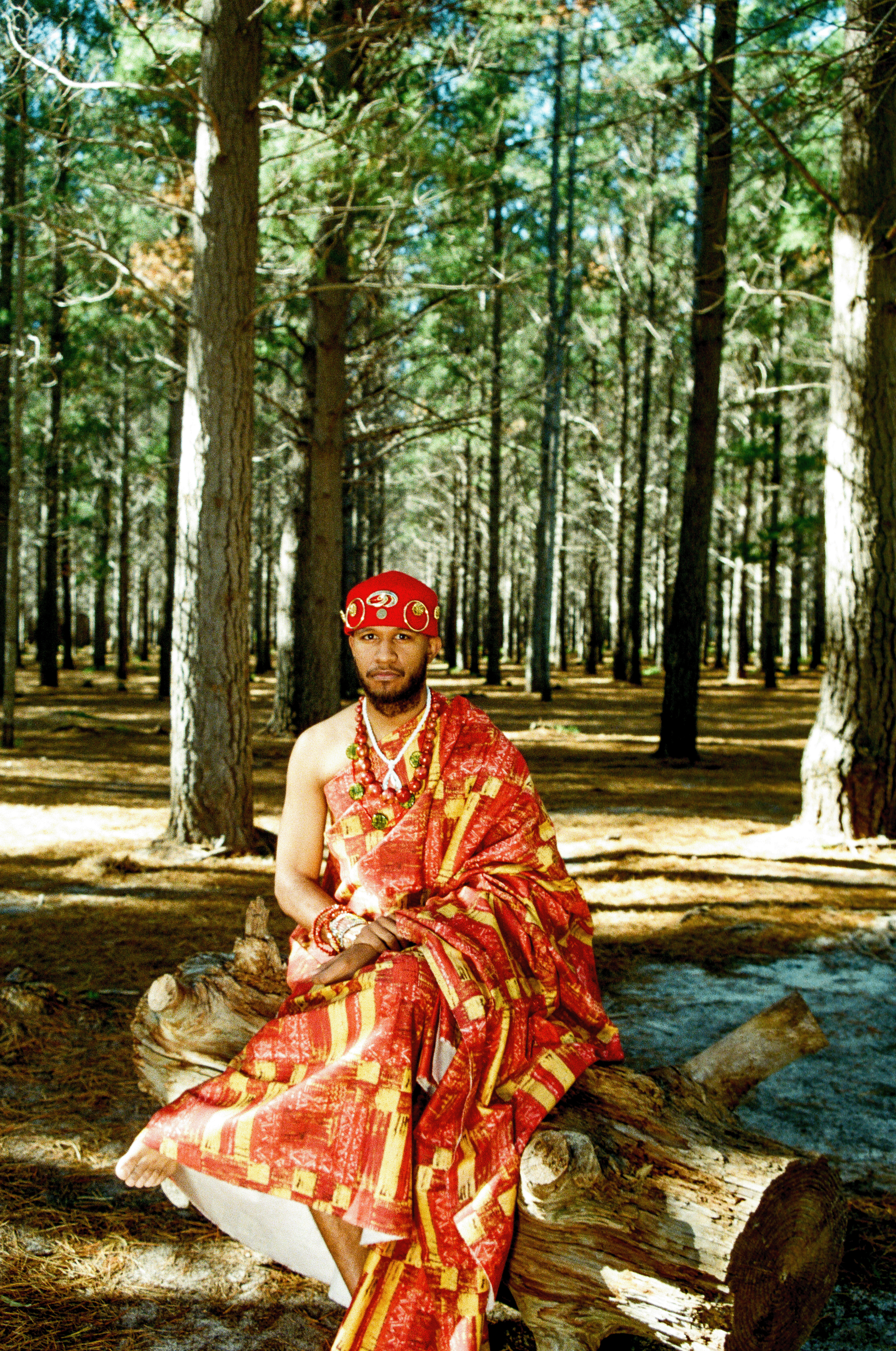 A man dressed in traditional African attire with red patterned fabric, jewelry, and a head wrap, sitting on a log in a forest.