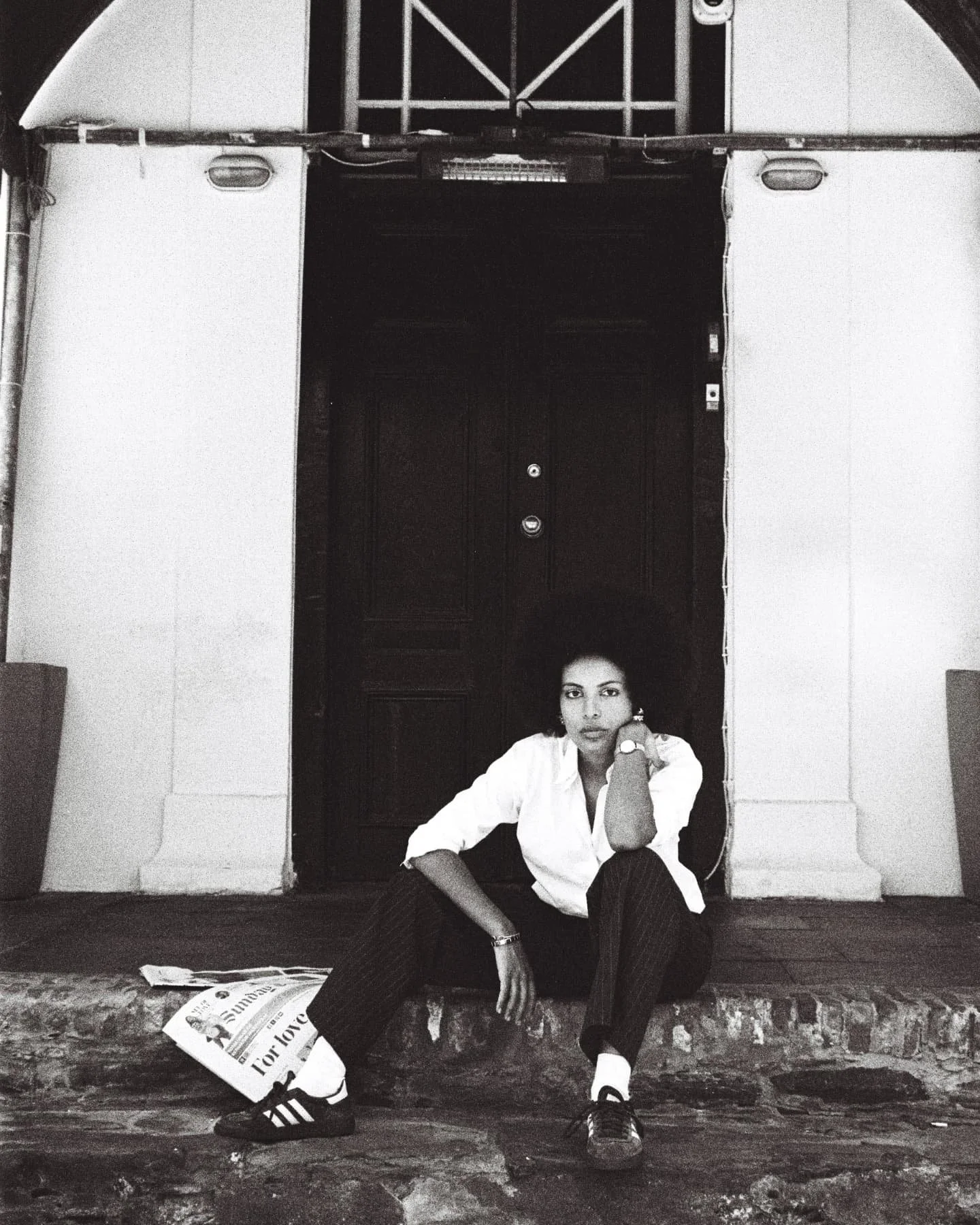 Mila Smith sitting on a stone doorstep in front of a dark door, resting her chin on her hand, with a newspaper on the ground.