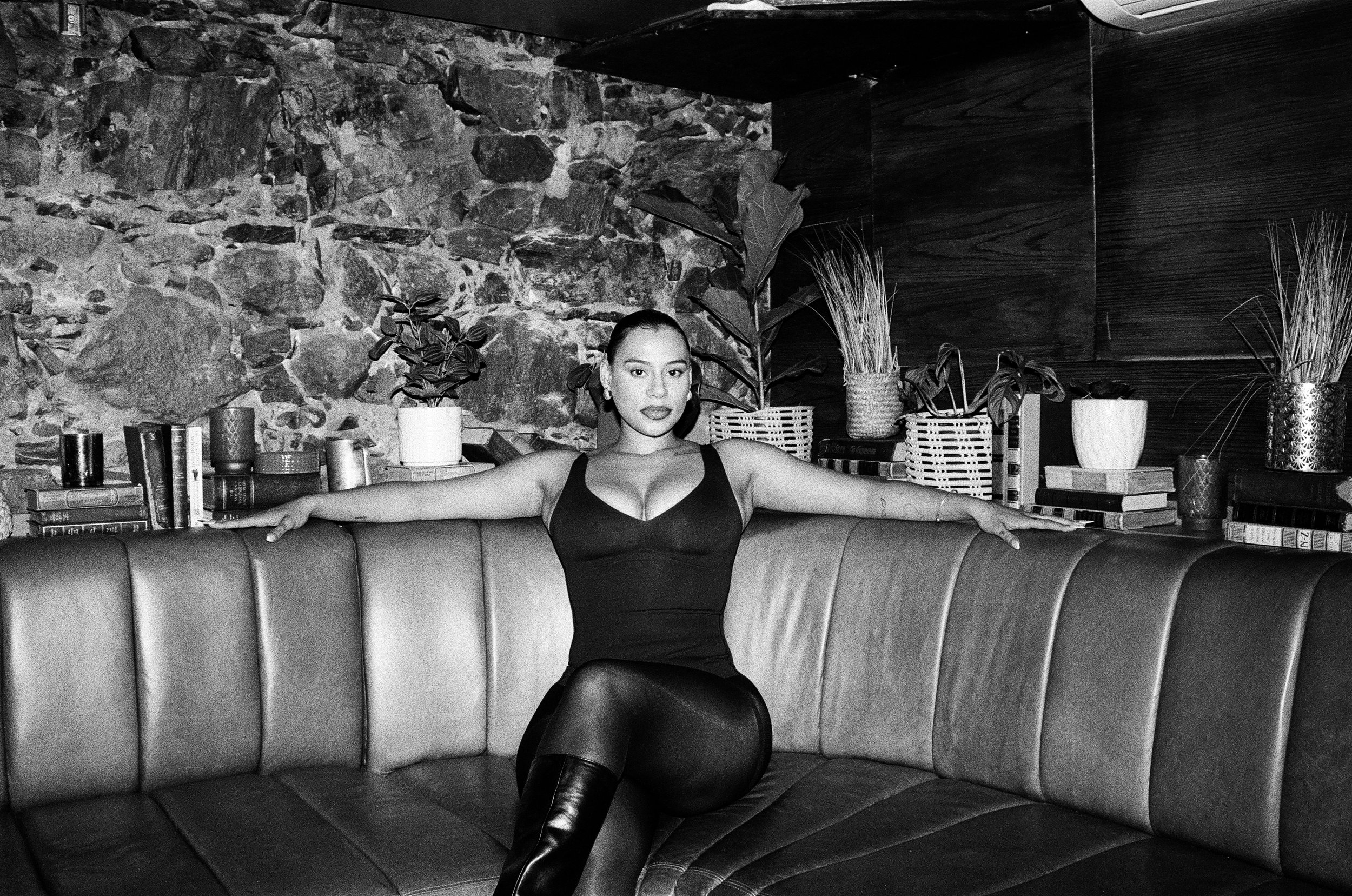 Kimberleigh Venty in a black dress and knee-high boots sitting on a curved sofa with her arms outstretched, in a room with a stone and wood interior, and plants and books on a shelf behind her.