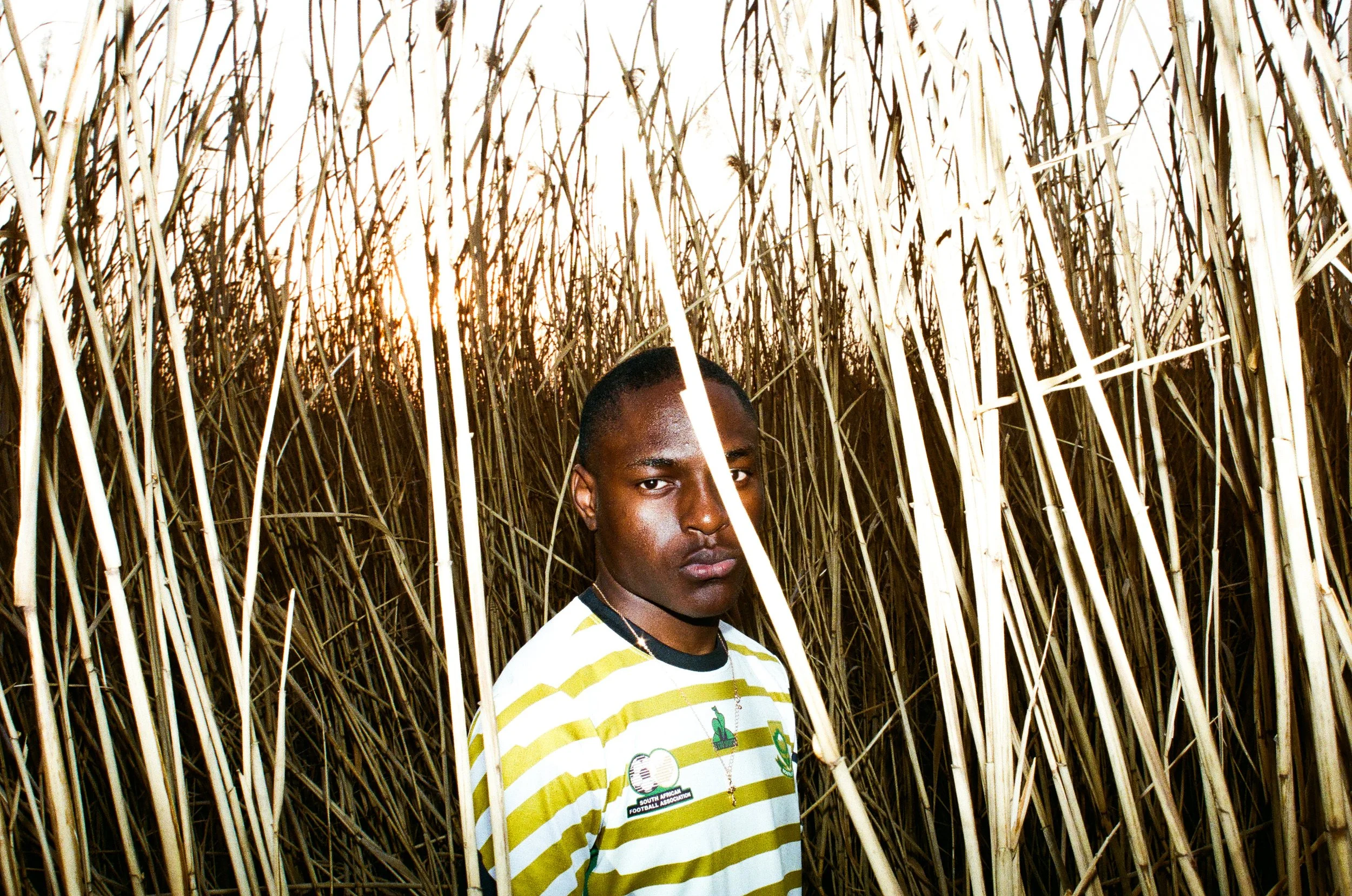 Young man standing in a field of tall, dry grass at sunset, wearing a striped shirt and a necklaces.