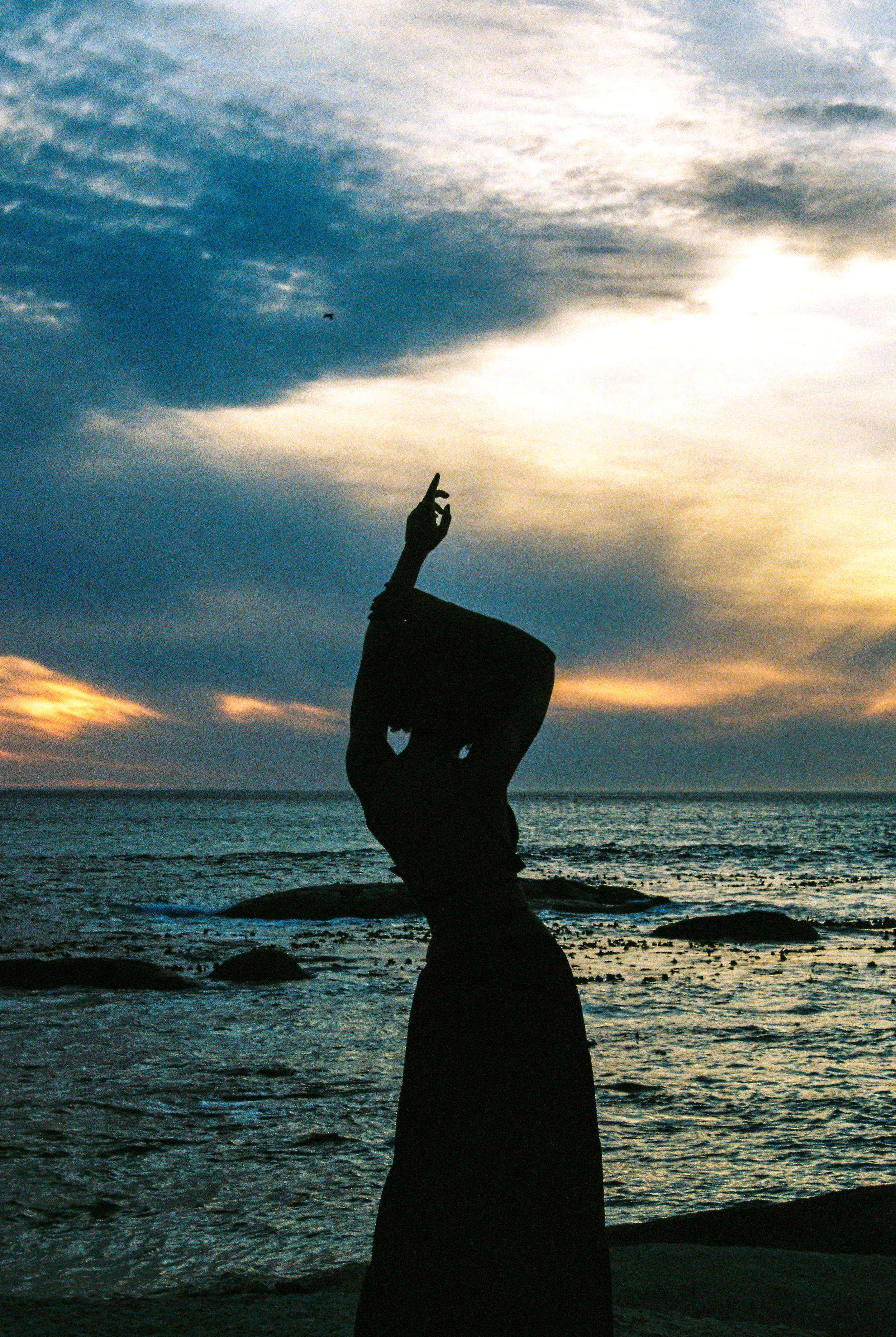 Silhouette of a person standing on rocks by the ocean at sunset, with one arm raised and fingers in a mudra pose.