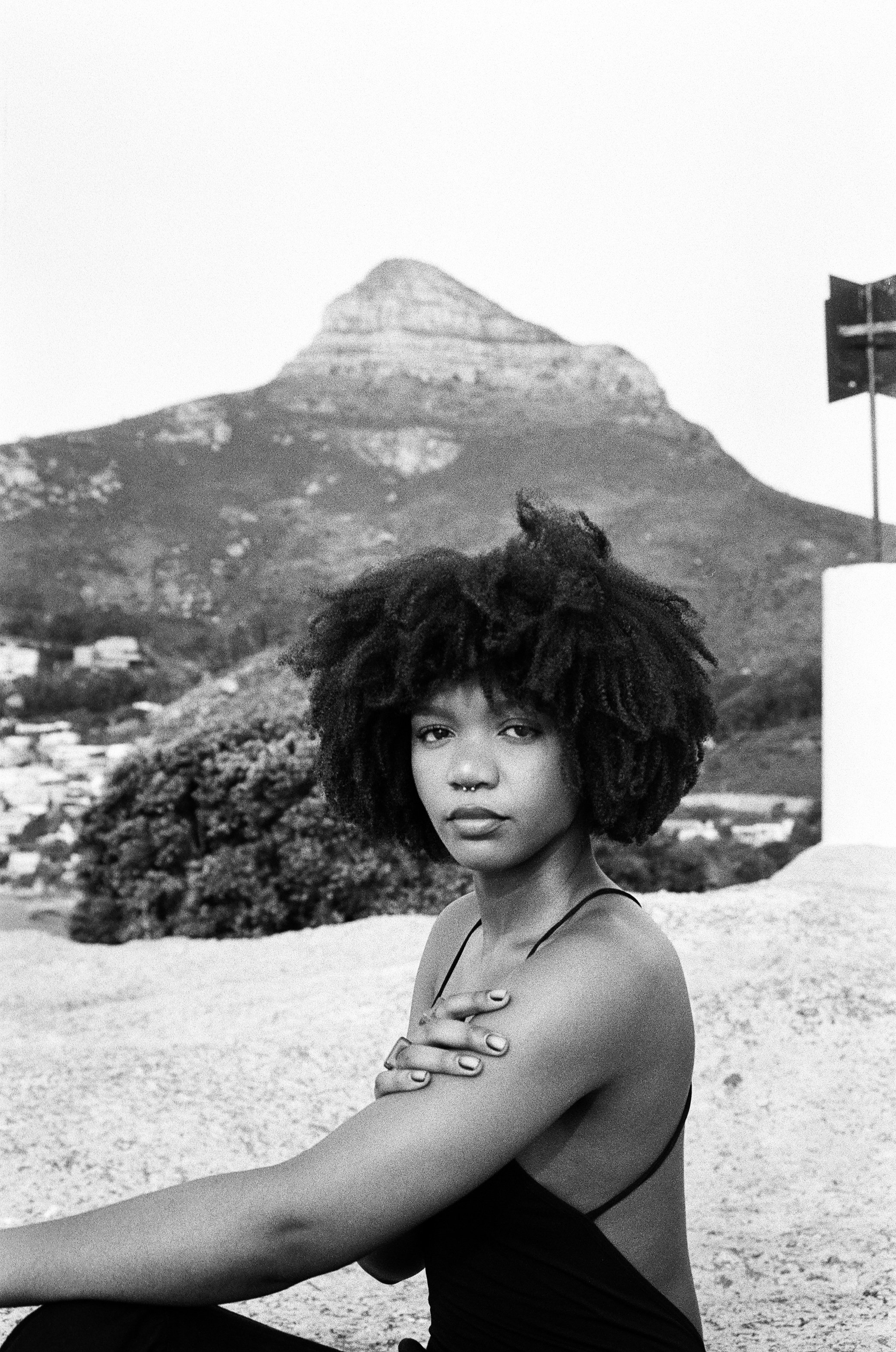 A young woman with natural curly hair and a septum piercing sitting outdoors, with a mountain and landscape in the background, in black and white.