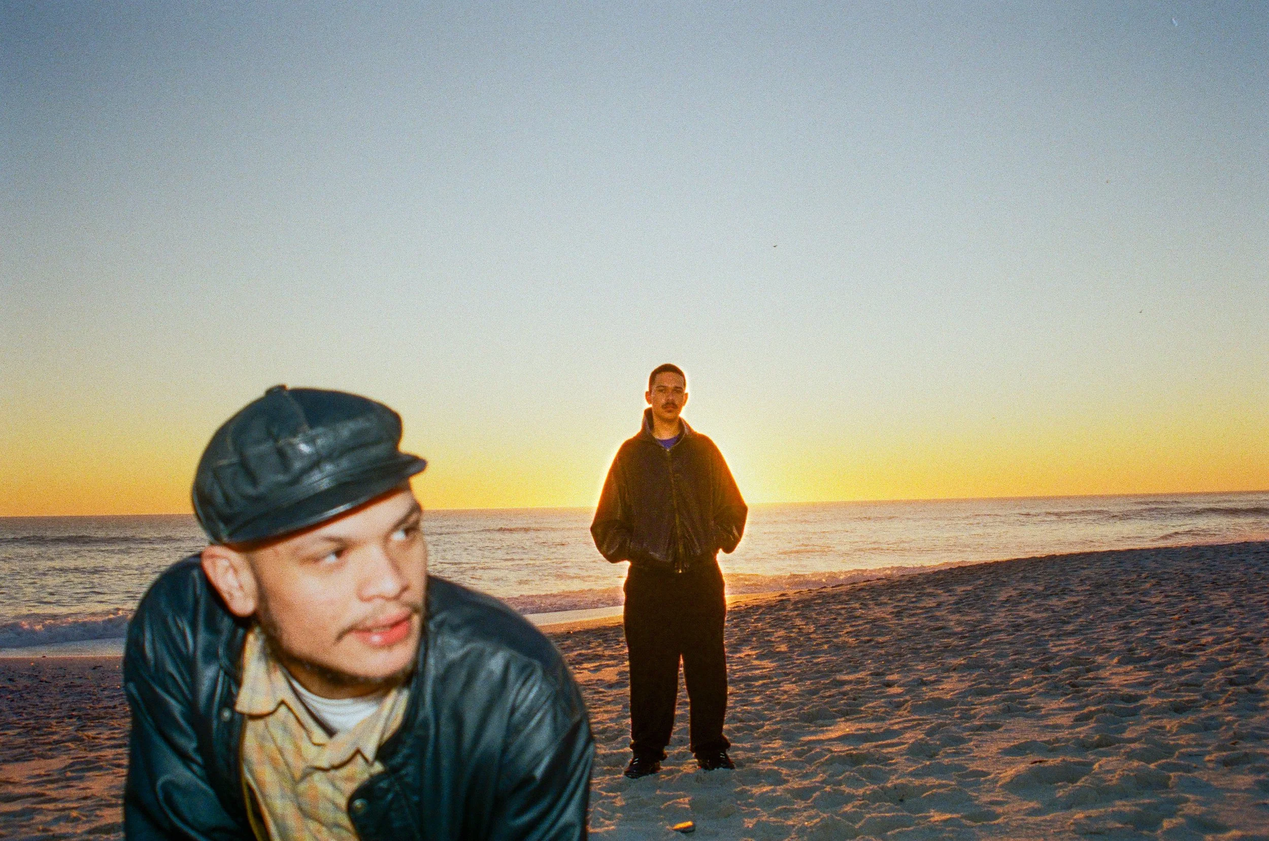 Jarry Pitboi and Permi Kwaali on the beach at sunset, one in a leather jacket in the foreground, the other in a dark hoodie in the background.