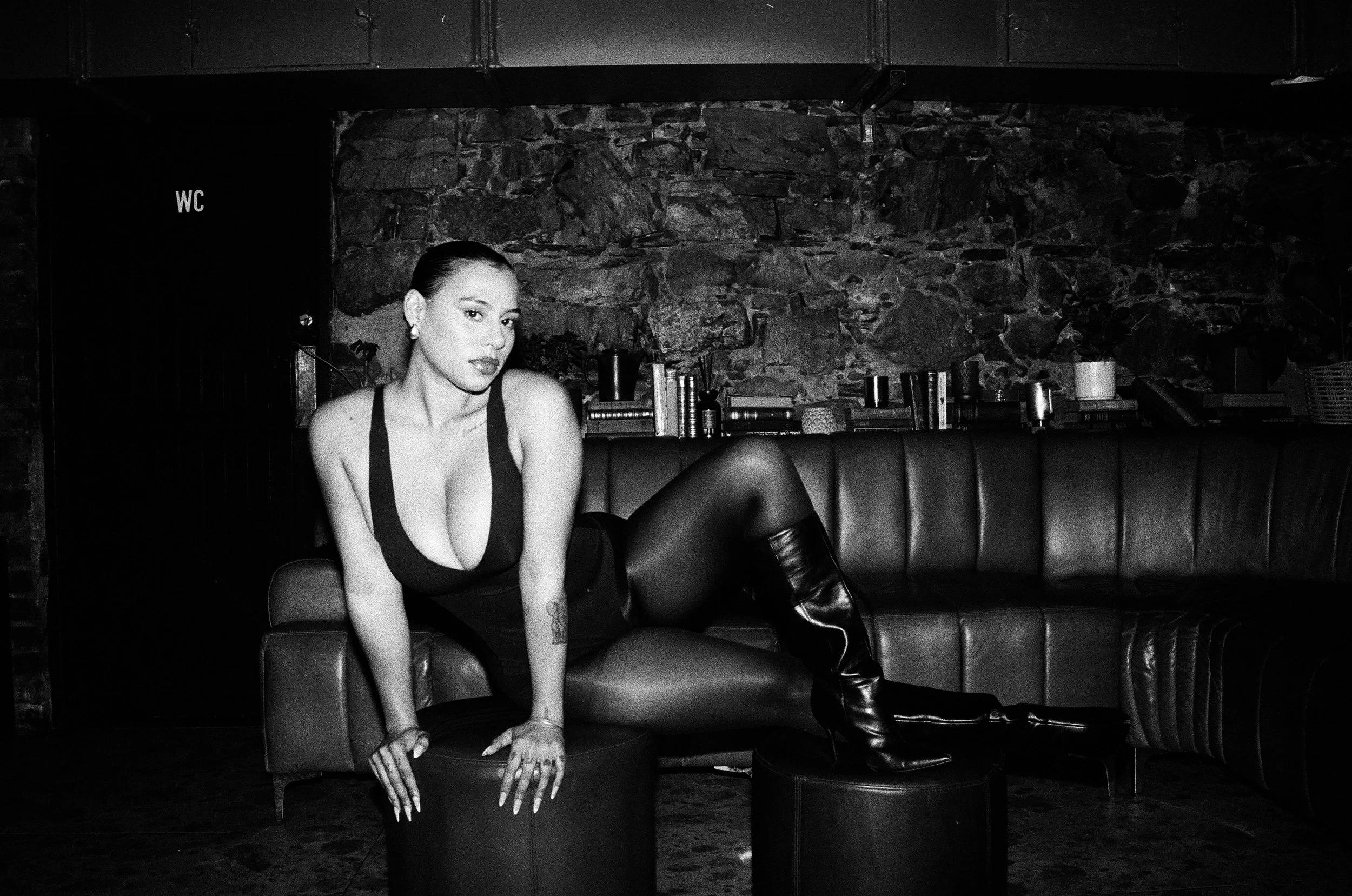 Kimberleigh Venty sitting on a stool with one leg raised on another stool in a dimly lit room with a stone wall background. She is wearing a sleeveless top, tights, and knee-high boots, with her hair slicked back.
