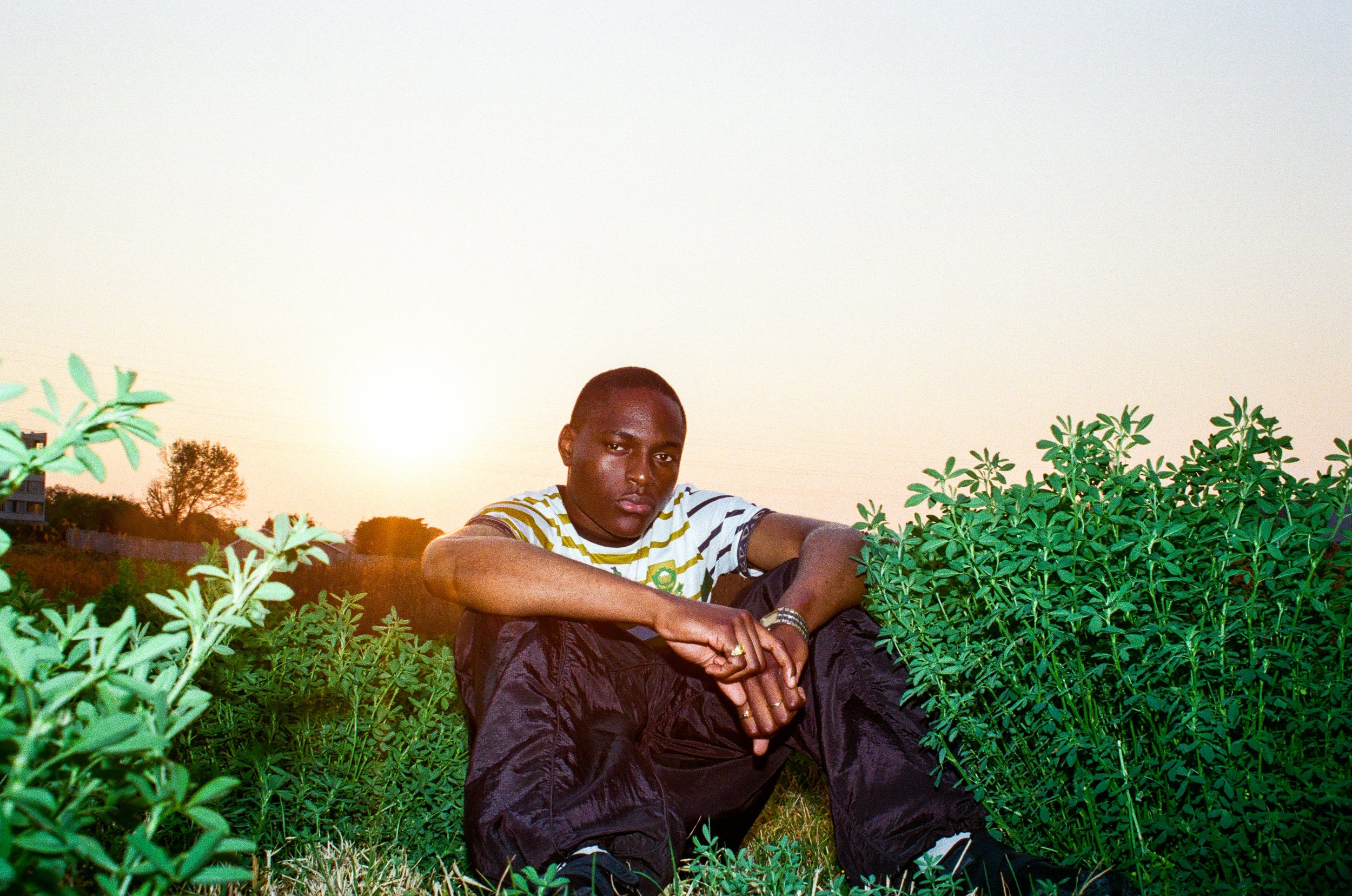 Joshua Futura sitting outdoors among green bushes at sunset, wearing a striped shirt and black pants, with a serious expression.