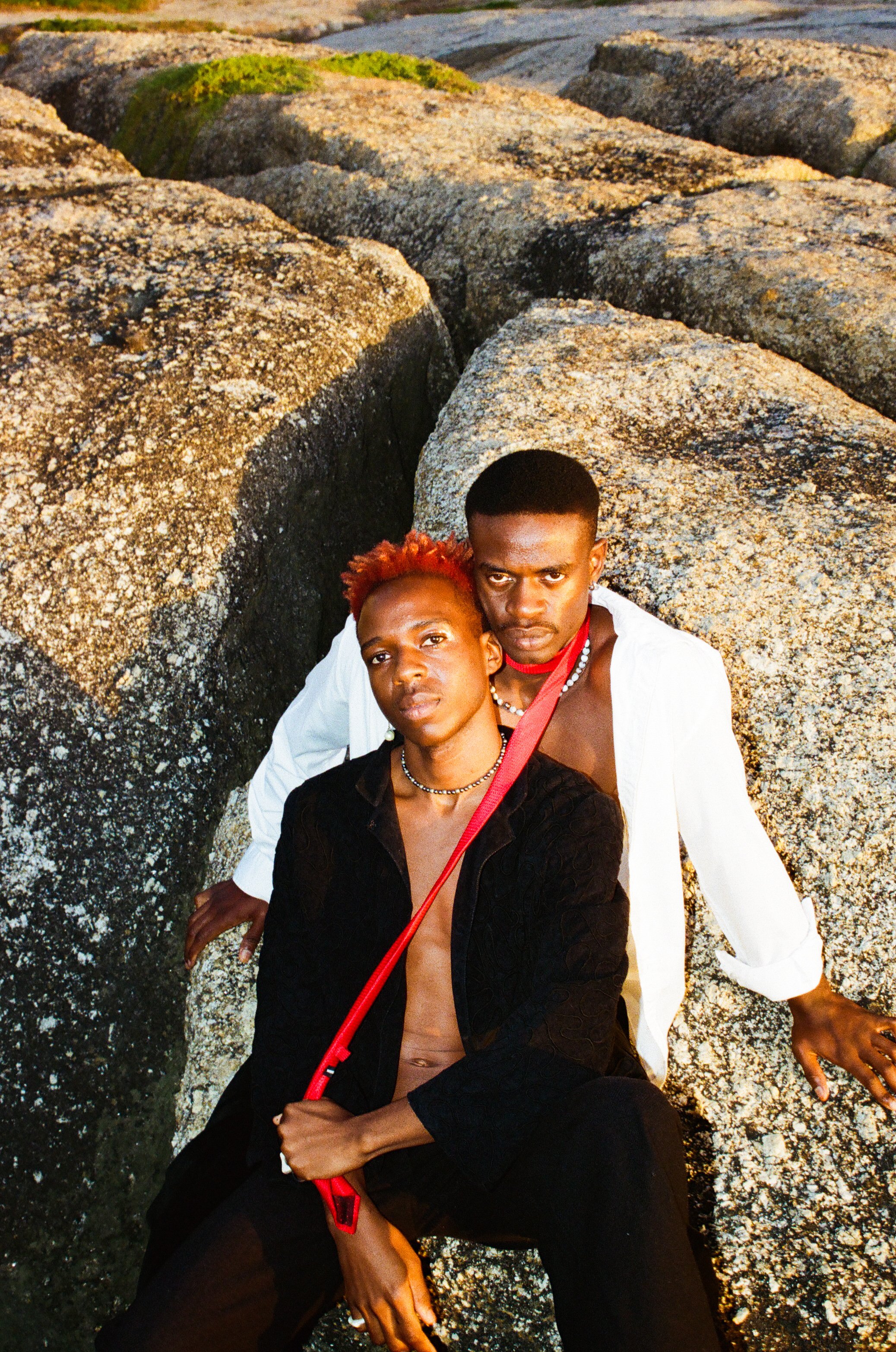 Dnemas and a friend sitting on rocks near a body of water, looking into the camera. One has red hair and is wearing a black jacket, the other has short hair and is wearing a white shirt.
