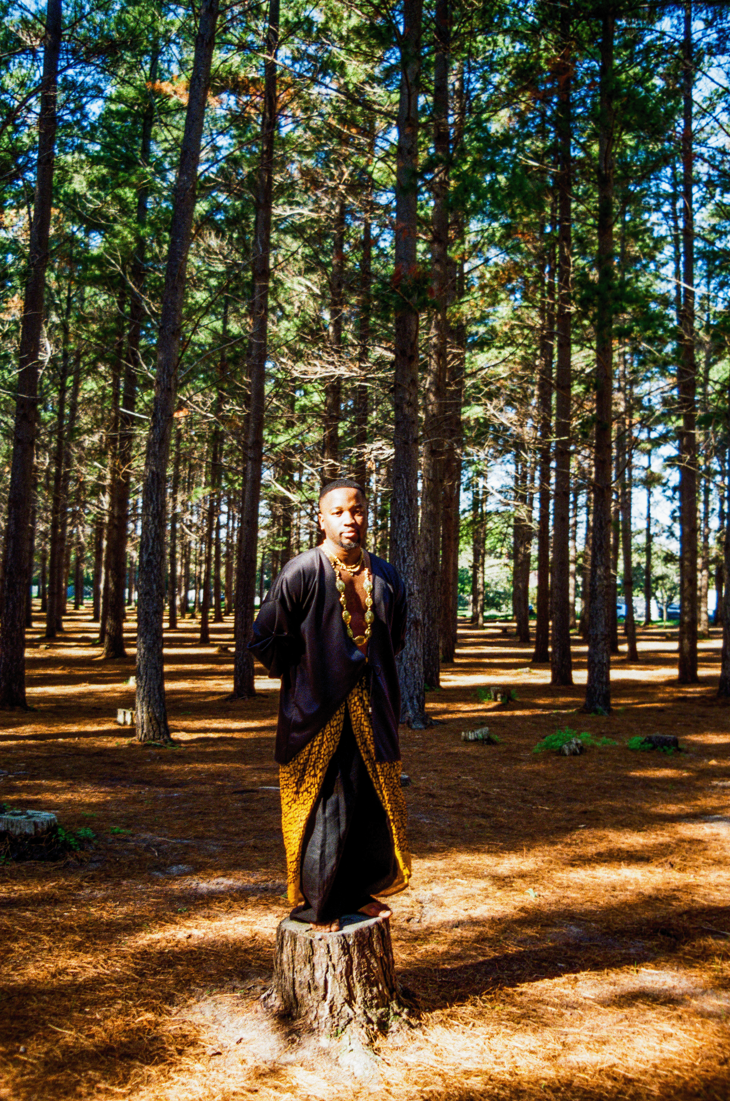 Owethu Ndwandwe dressed in traditional African clothing with gold jewelry, standing on a tree stump in a forest with tall pine trees and a blue sky.